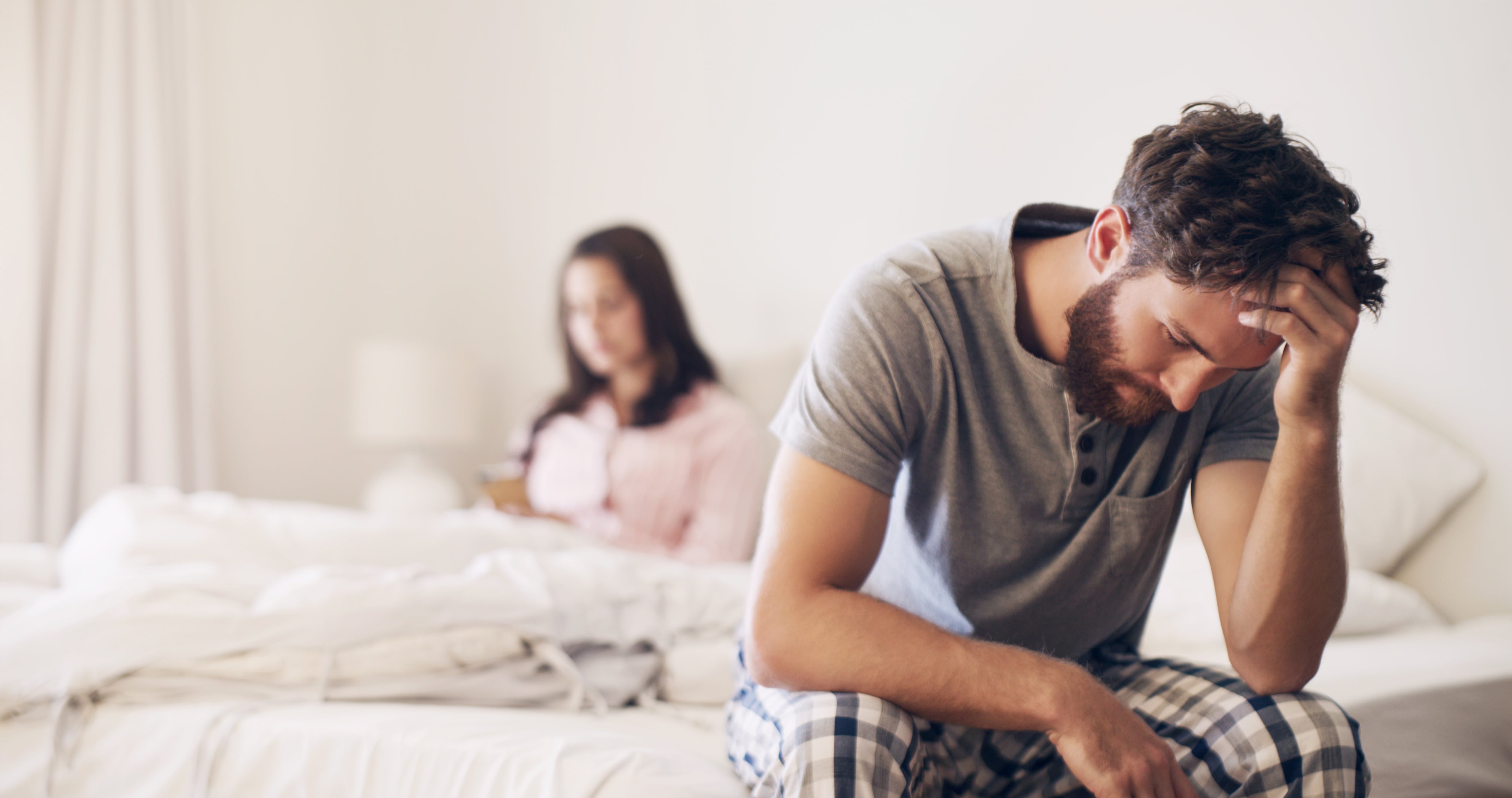 A man on the edge of one side of the bed with his head in his hands appearing frustrated. The woman is in the background of the image sitting under the covers. 