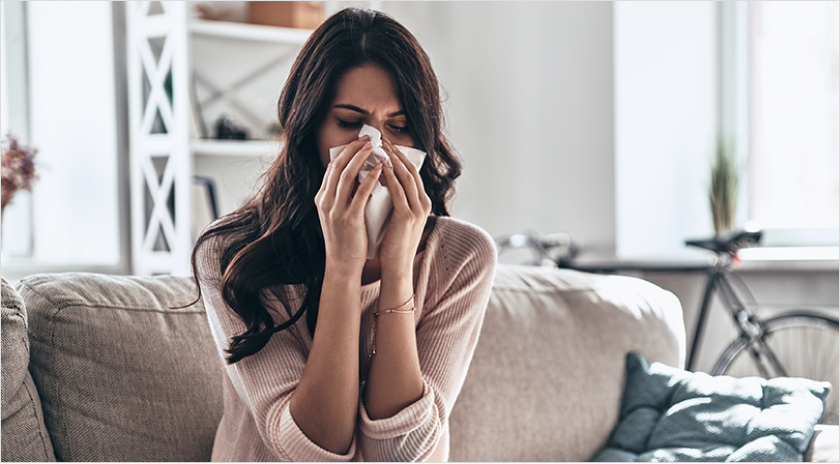 Woman sneezing into tissue while sitting on a sofa inside her home