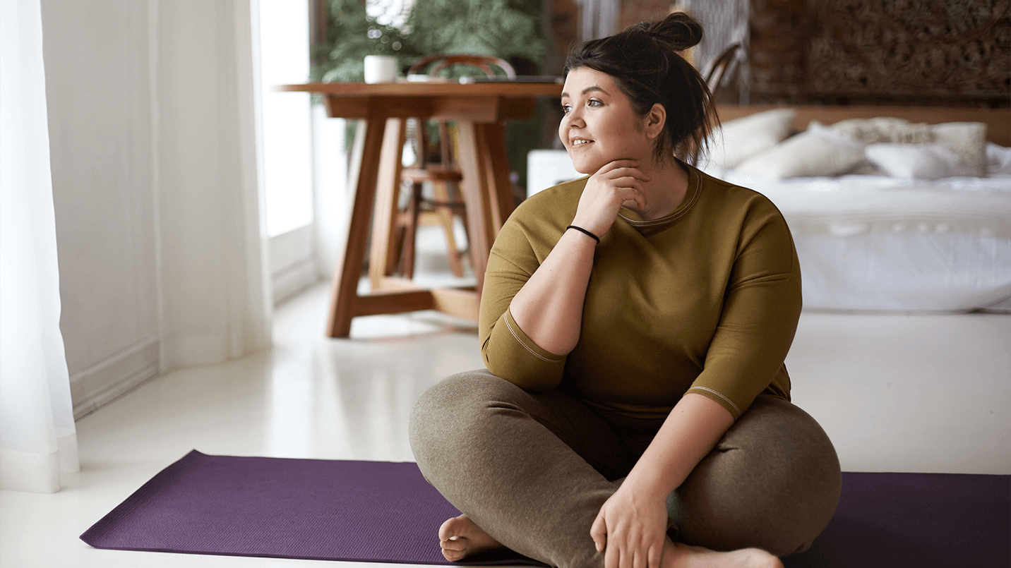 overweight woman smiling and sitting on yoga mat looking out a window