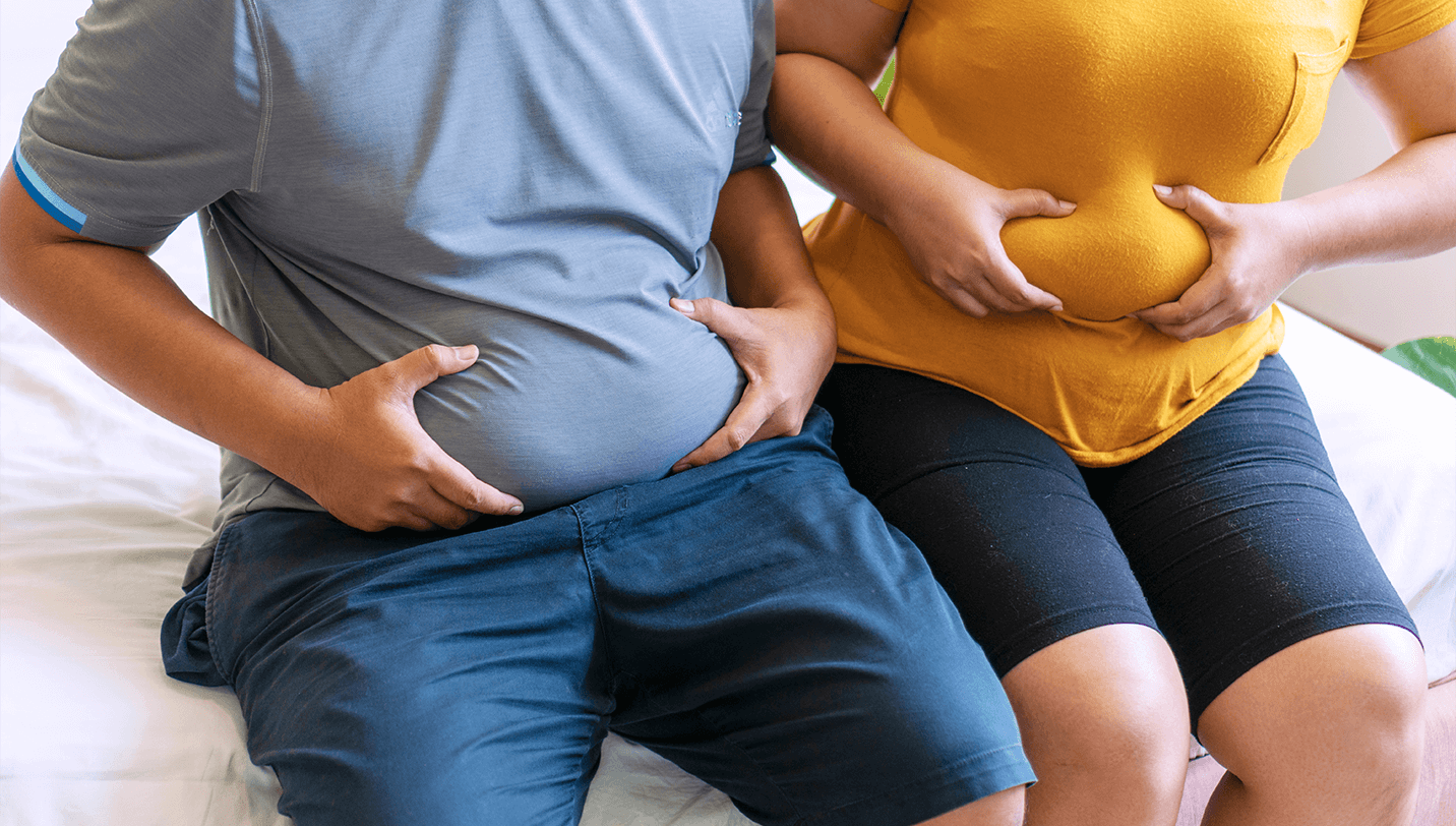 A man and woman sitting on a bed pinching their stomach fat