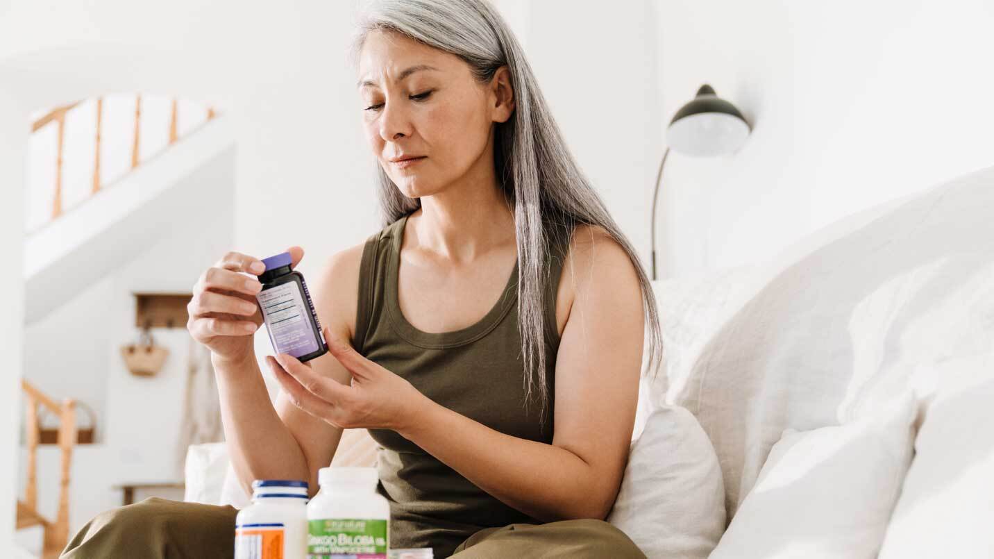 women with grey hair looking at vitamin bottle and sitting on white couch