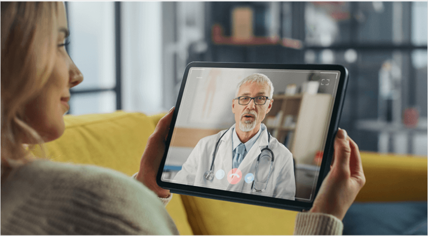 A woman sitting on her sofa holding a tablet that shows a male doctor virtually chatting with her.