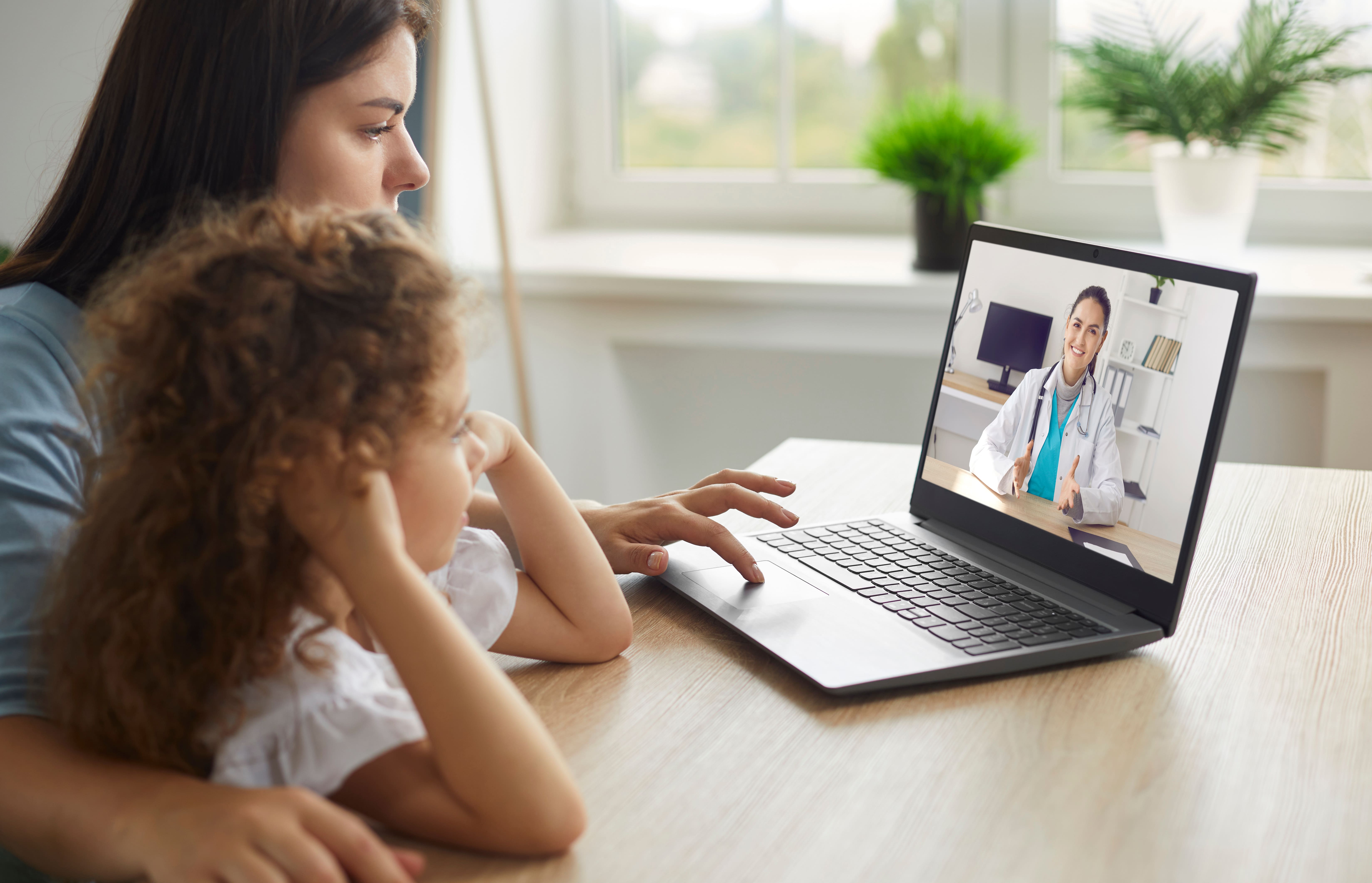 A mother and daughter sitting at a table looking a computer screen undergoing a telehealth session with a doctor. 