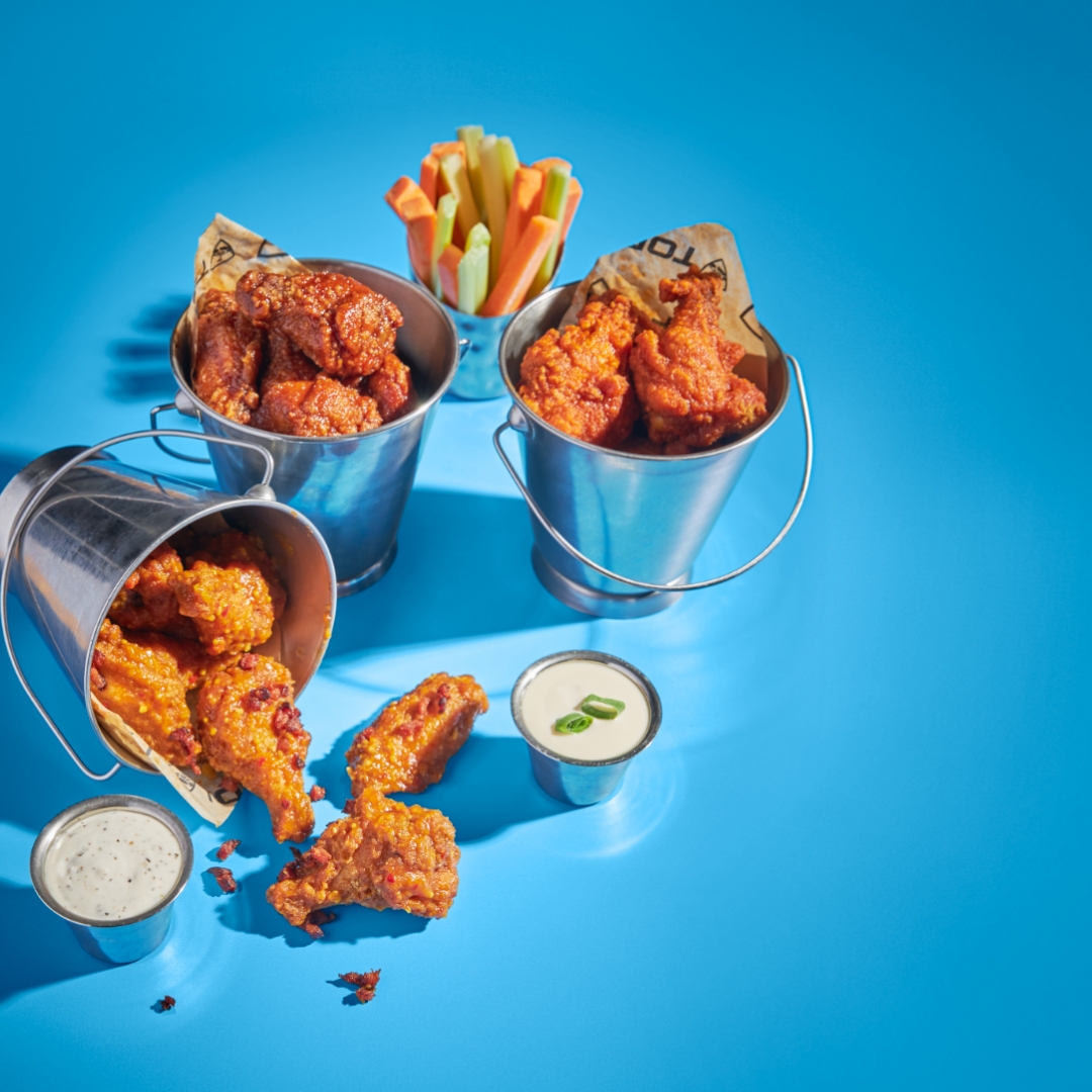 Three metal buckets filled with buffalo chicken wings and carrot and celery sticks are arranged on a blue surface, with two cups of ranch dipping sauce nearby.