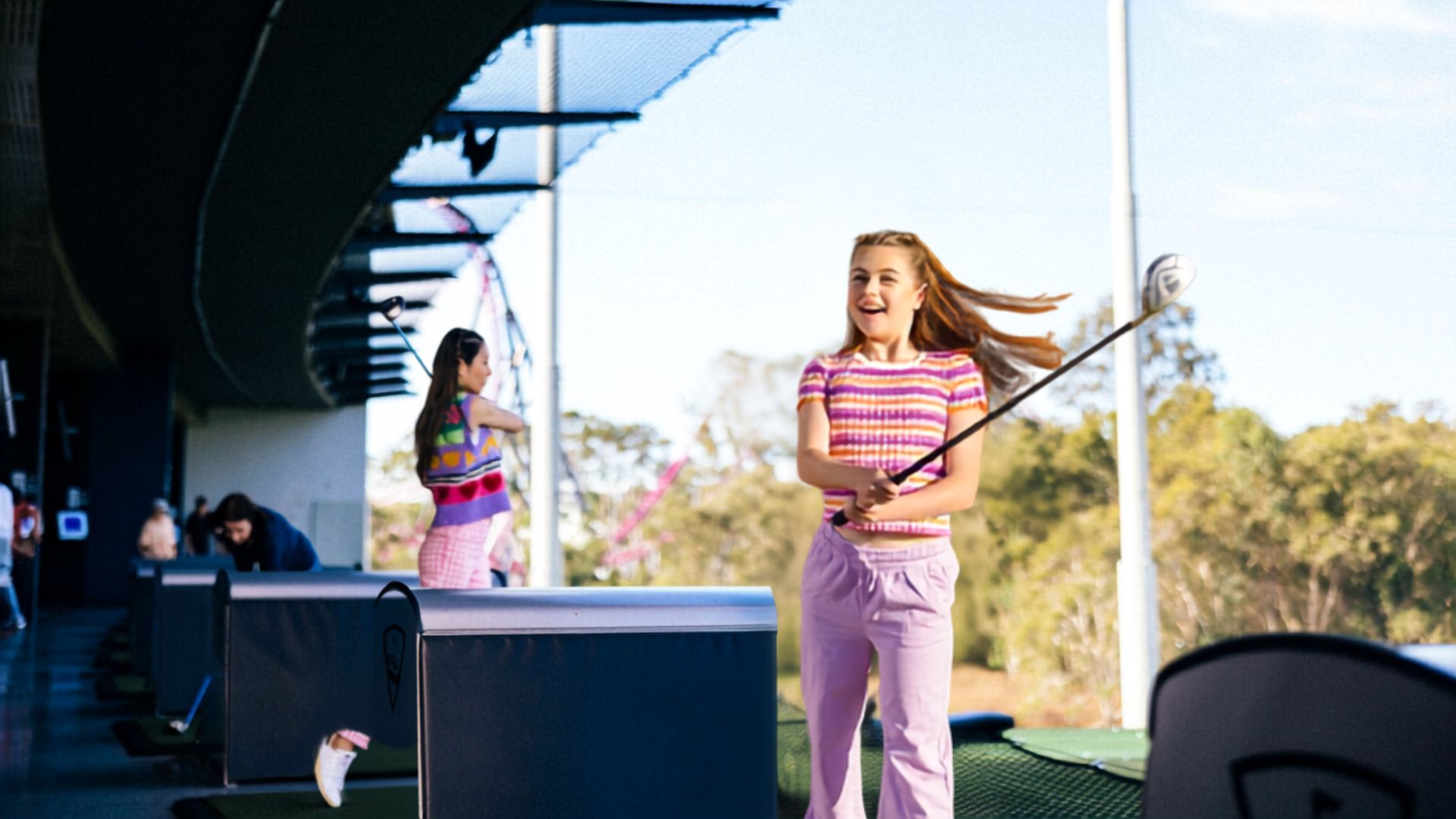 Two young women practice their golf swings at a driving range. One smiles while swinging a club, and another stands in the background. Both wear colorful clothing, and the area is surrounded by trees.
