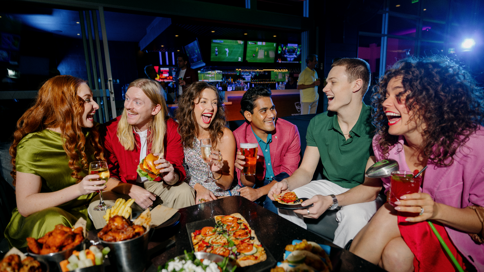 Six friends sit together at a table, laughing and enjoying drinks and food at a lively indoor venue with sports playing on big screens in the background.