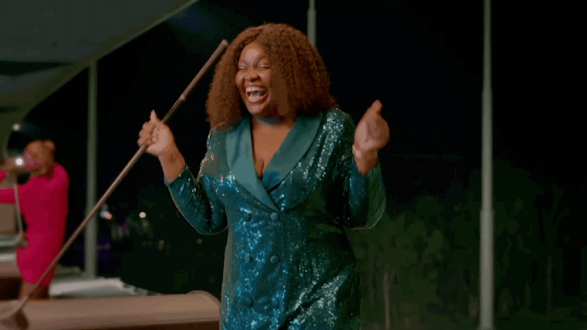 A woman in a sparkly green dress laughs joyfully and dances with excitement, while holding a pool cue. Another person in pink stands in the background.