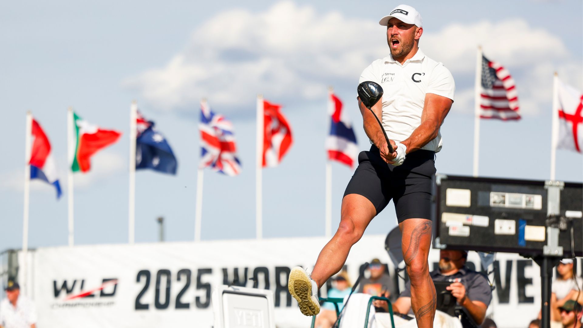 A man in athletic wear swings a golf club powerfully, mid-air, at a competition with multiple international flags and a "2025 World" event banner visible in the background.