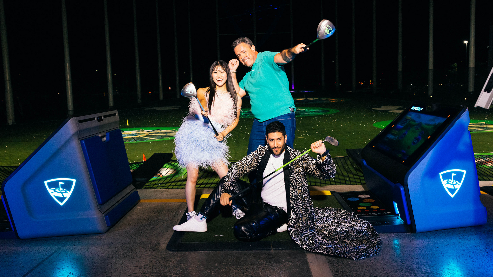 Three people pose with golf clubs at an indoor driving range. Two stand smiling, while one kneels playfully in a patterned coat. Bright lights and golf targets are visible in the background.