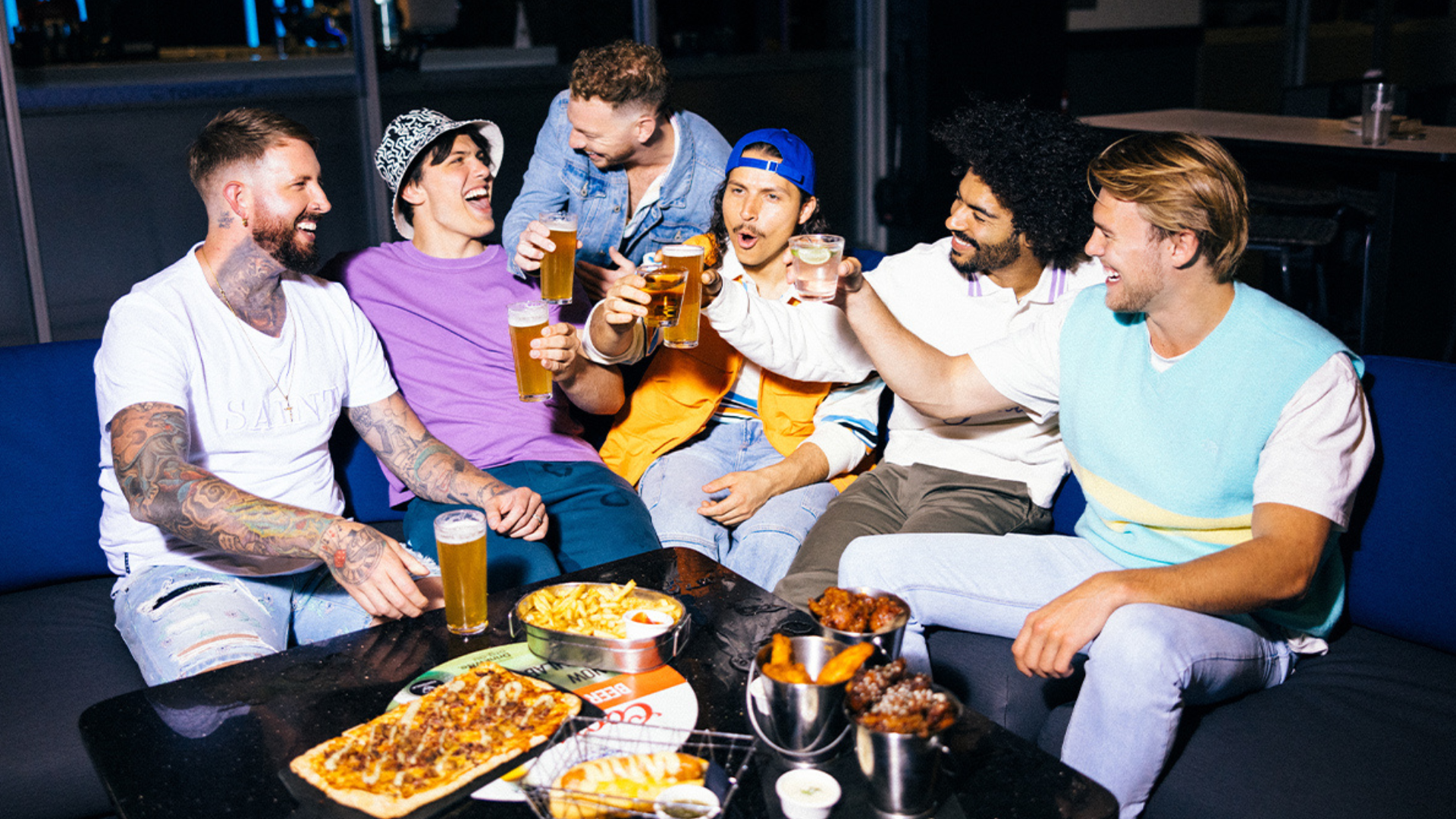 Six men sit on a couch, laughing and toasting with beer glasses. Food like wings, fries, and pizza is on the table in front of them, suggesting they are enjoying a casual, fun night out together.