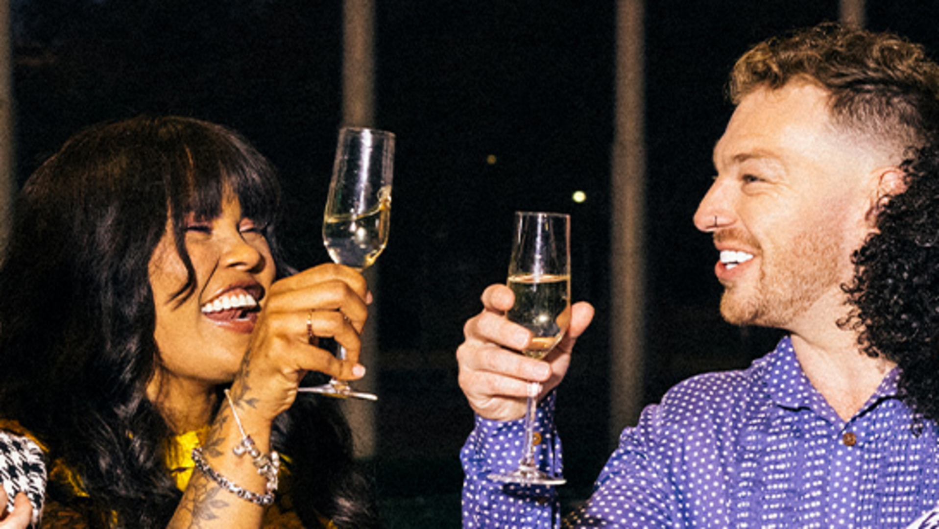 Two people smiling and raising champagne glasses in a toast, appearing to celebrate together in a festive setting.