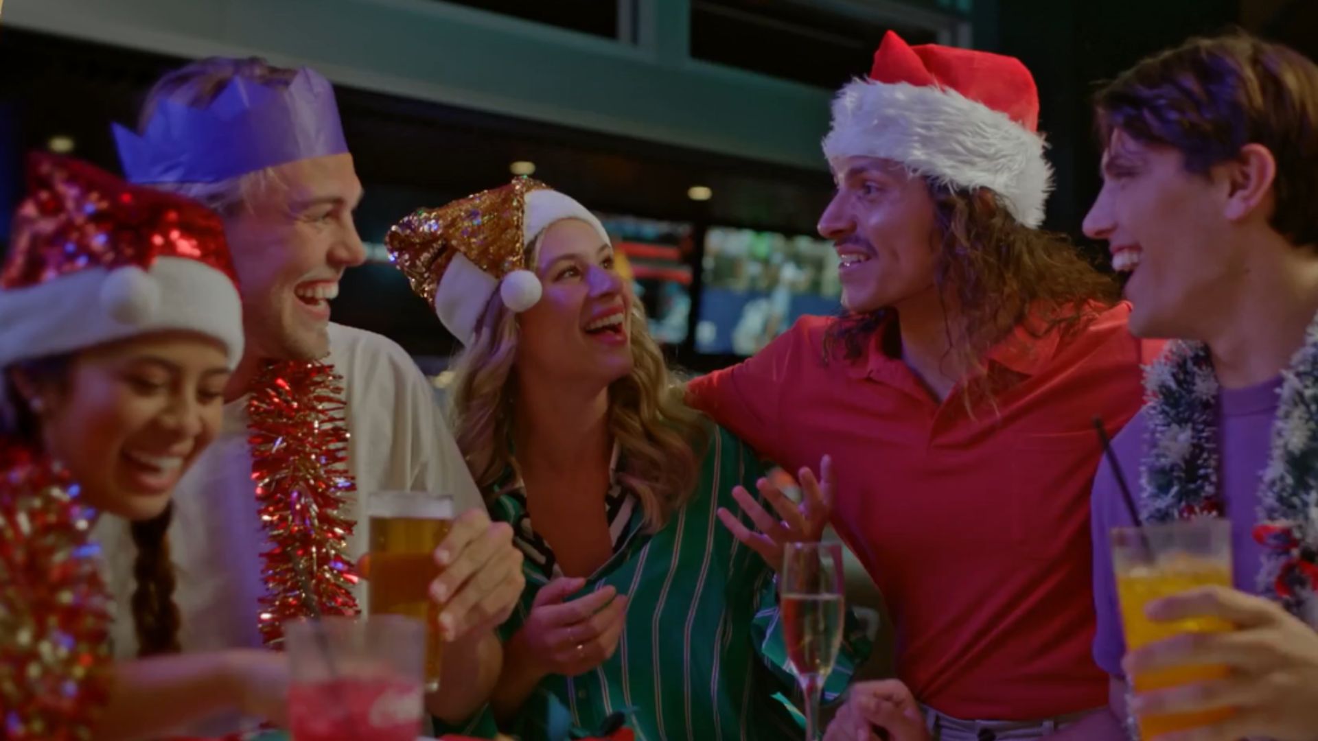 Five people in festive hats and tinsel smile and laugh together at a bar, holding drinks, celebrating in a lively, cheerful atmosphere.