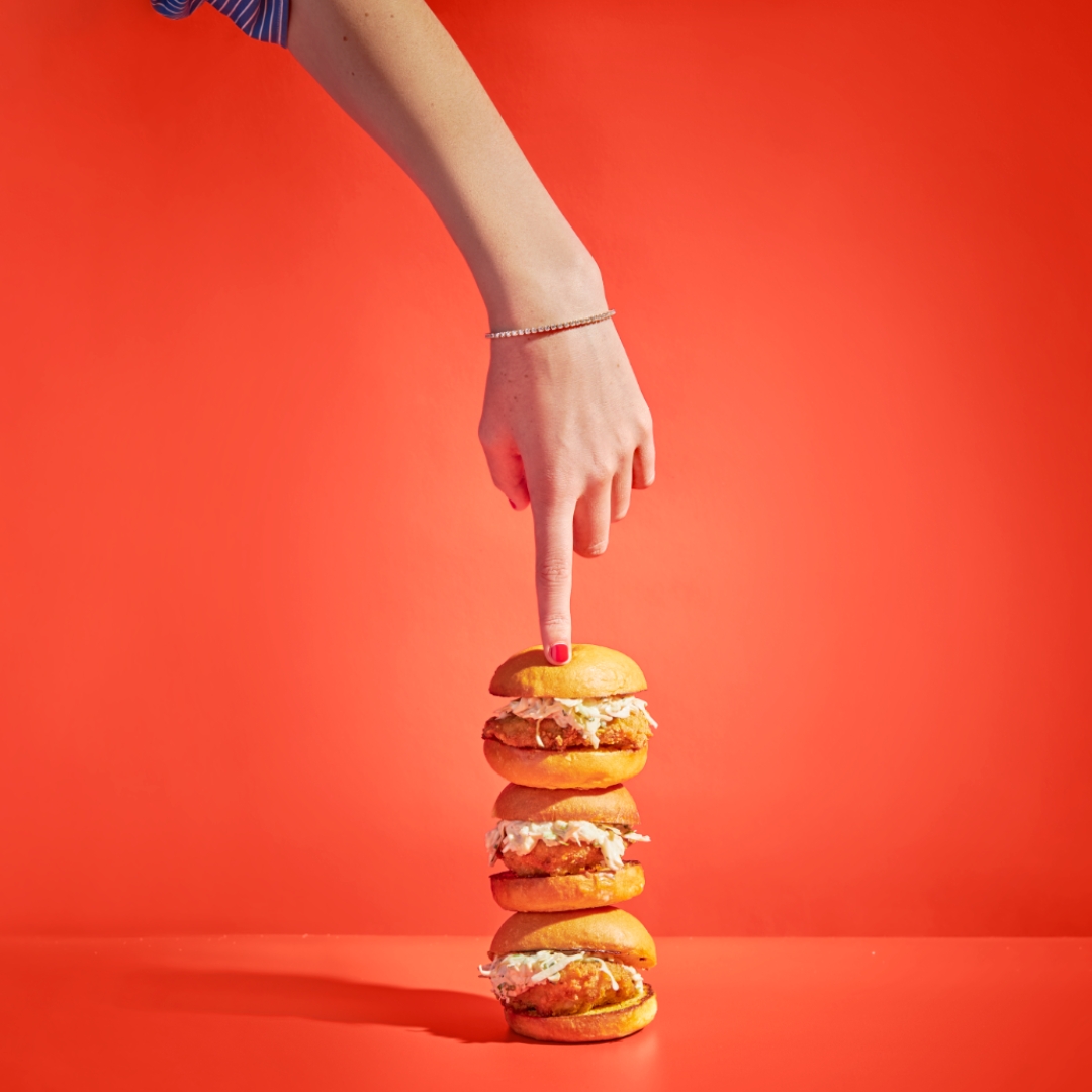 A hand with red nail polish balances four stacked chicken sandwiches against a bright red background.