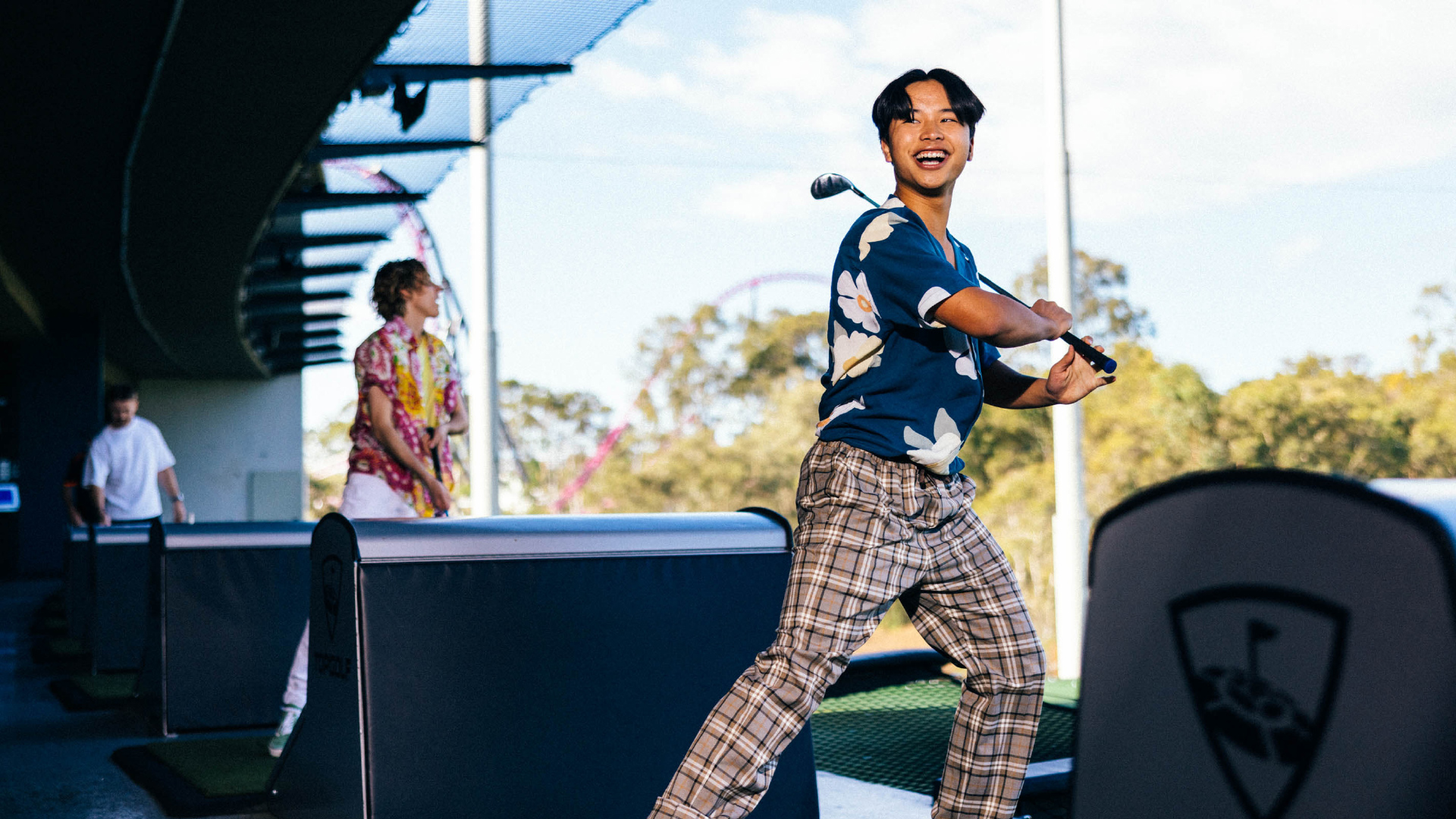 A smiling person in a blue floral shirt and plaid pants swings a golf club at a driving range, while others play in the background under a sunny sky.