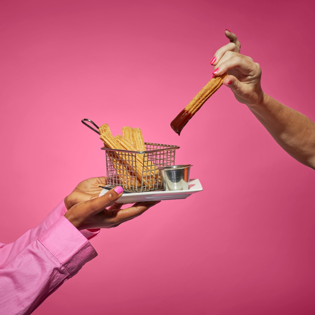 A person holds a basket of churros and a small cup of chocolate sauce while another hand dips a churro into the sauce, both against a bright pink background.