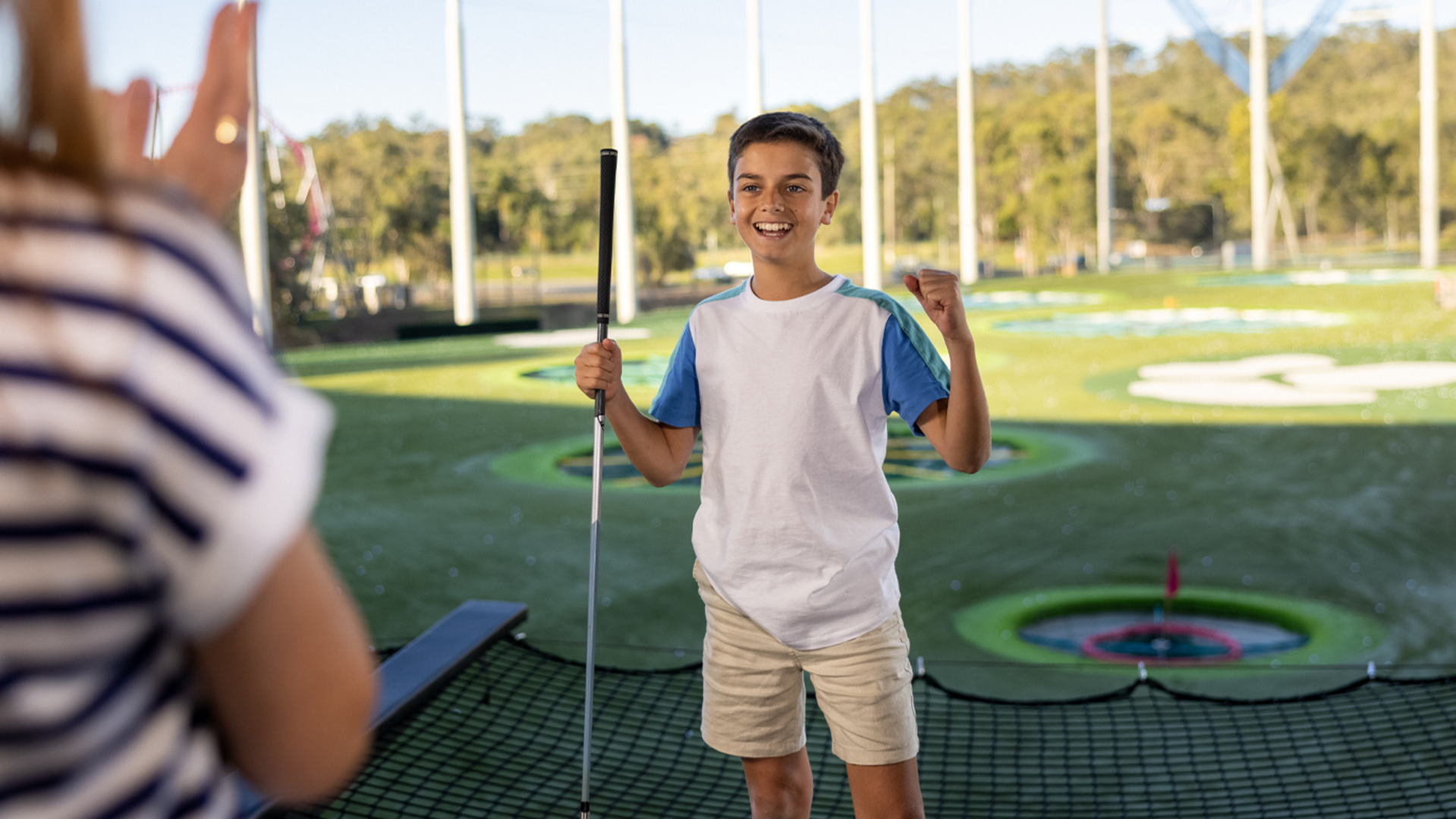 A smiling boy holding a golf club celebrates as he stands on a driving range, with colorful targets on the grass behind him and another person clapping in the foreground.