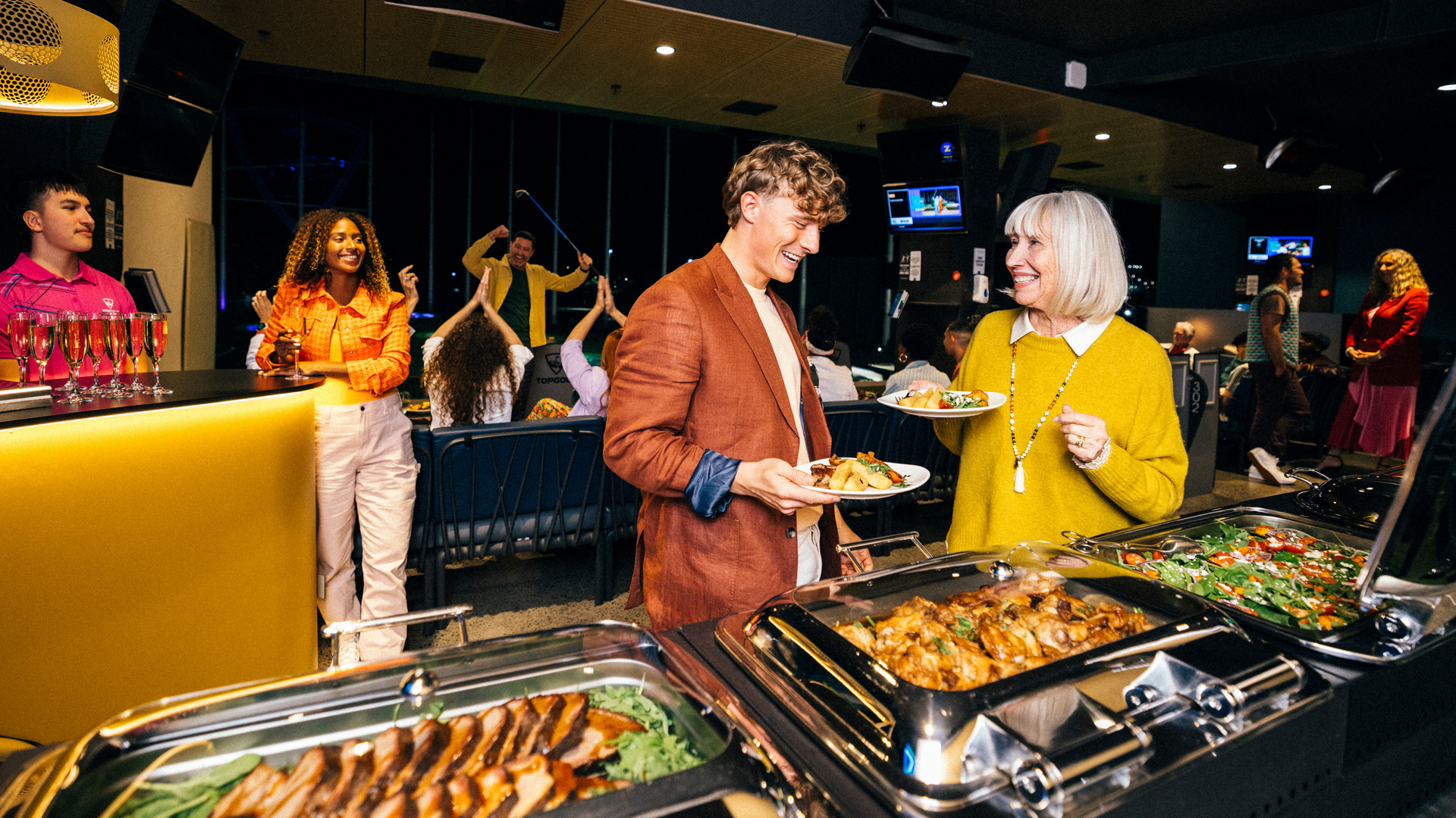 Two people stand by a buffet table serving themselves food at a lively indoor event, with others in the background dancing and socializing. The atmosphere is festive, with colorful lights and a variety of dishes displayed.