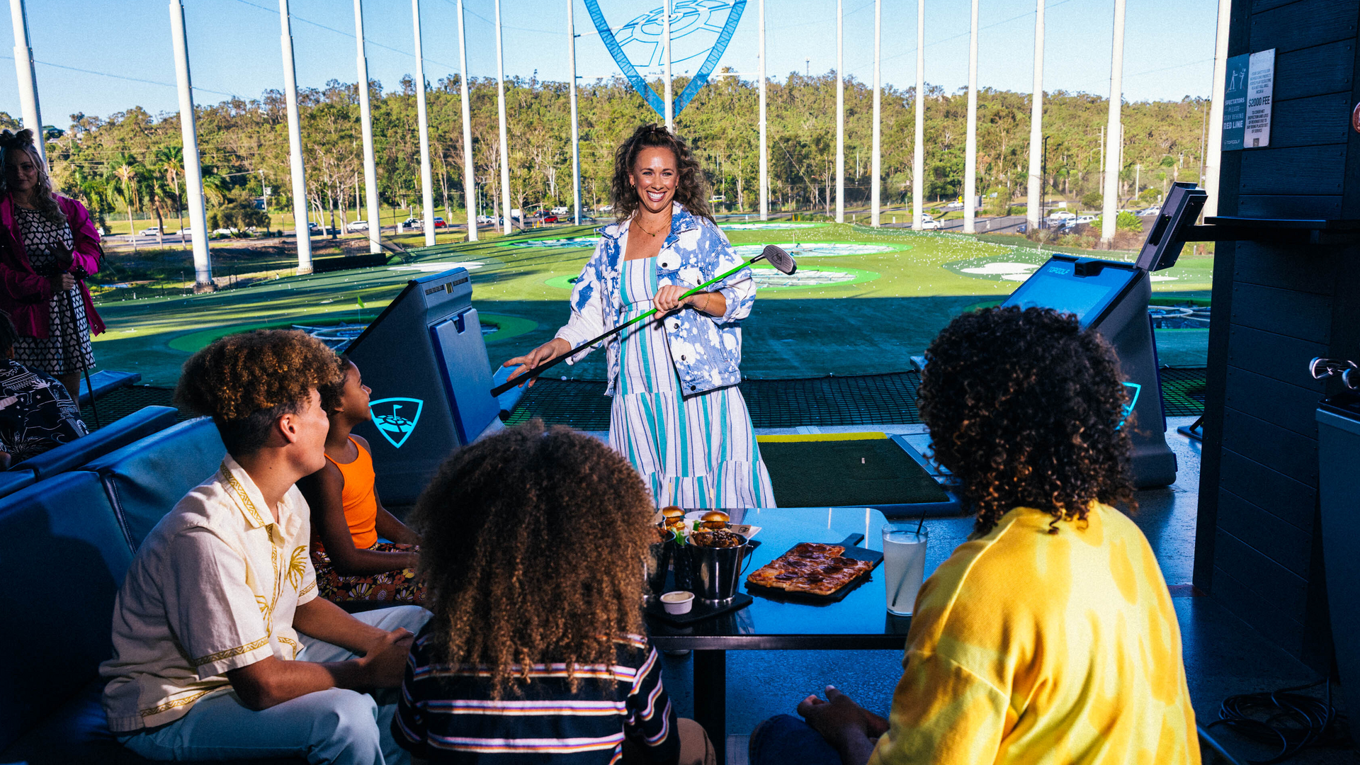A smiling woman stands with golf clubs at an outdoor driving range, while three people sit at a table with food and drinks, enjoying the sunny day together.