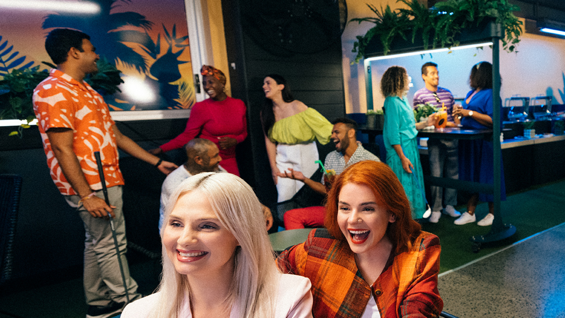 A group of people at a lively indoor party, smiling and laughing in colorful outfits. Two women are seated in the foreground, while others stand and chat near a buffet in the background.