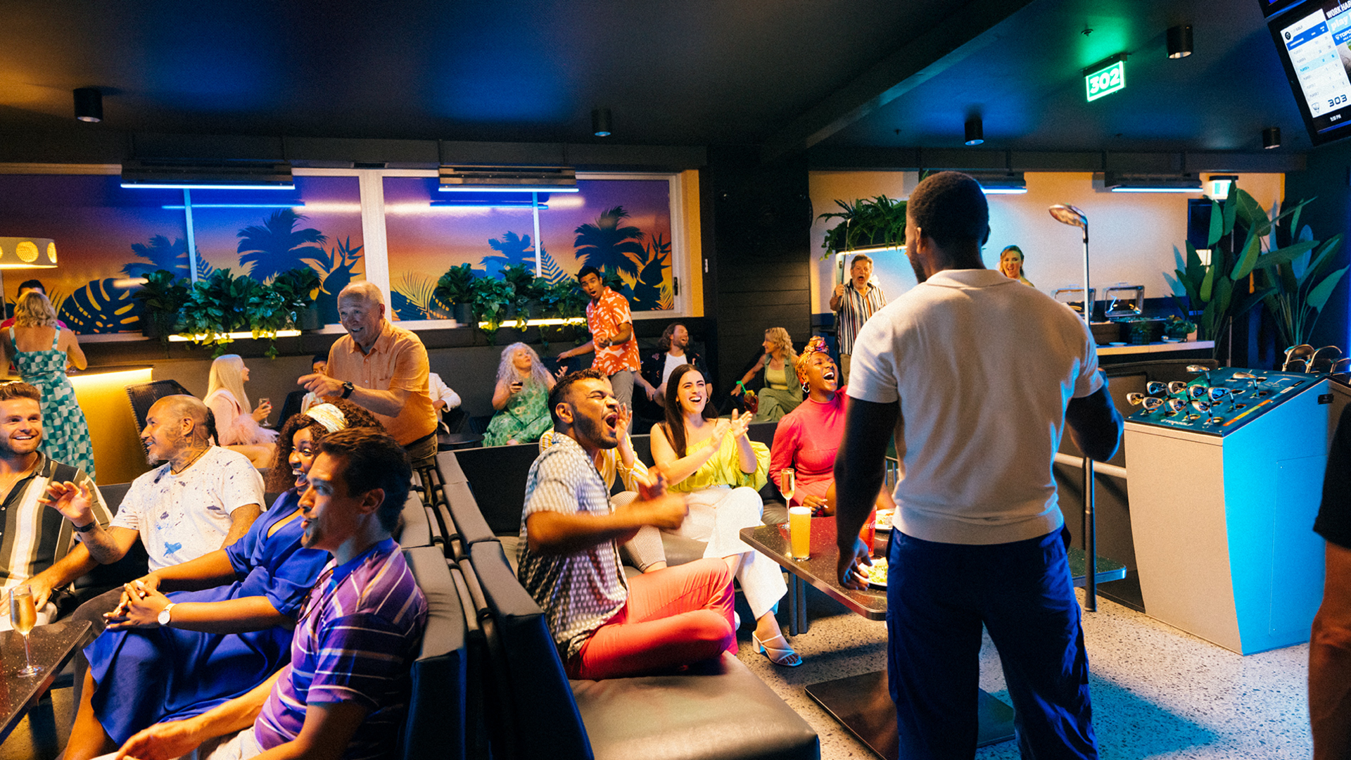 A group of people sit and laugh in a lively, colorful lounge or bar. One man stands facing the crowd, possibly performing or entertaining. Neon lights and tropical decor create a vibrant atmosphere.