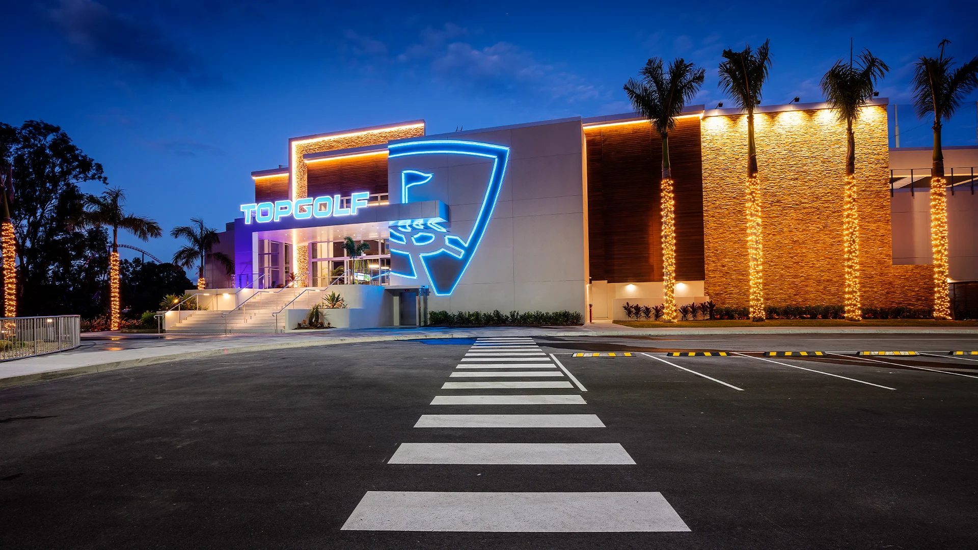 A Topgolf building with a neon sign and palm trees, offering exciting entertainment and competitive golf games. Visit us for pricing info and check our opening hours.