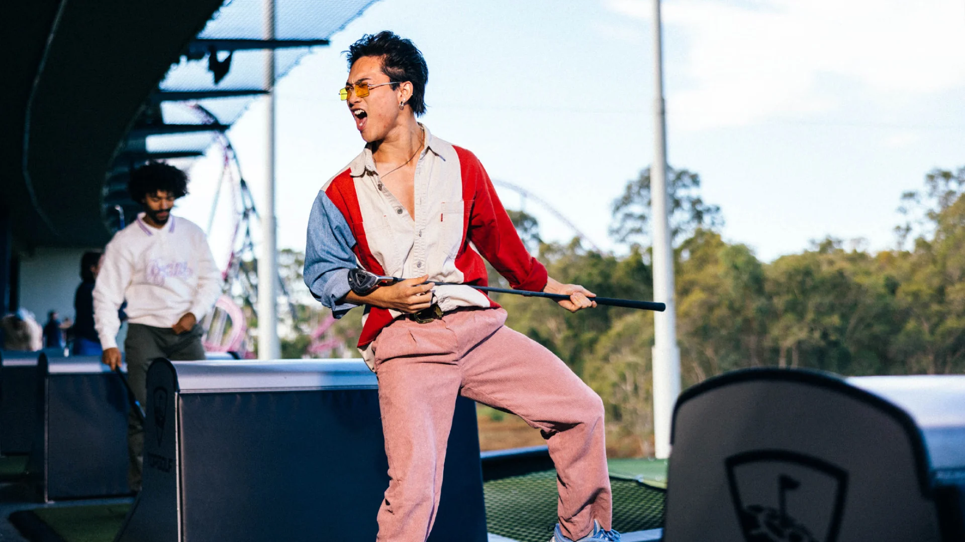 Person holding a golf club in a lively stance at a driving range, wearing a colorful outfit and sunglasses. Another person is in the background, with trees and a partially cloudy sky visible.