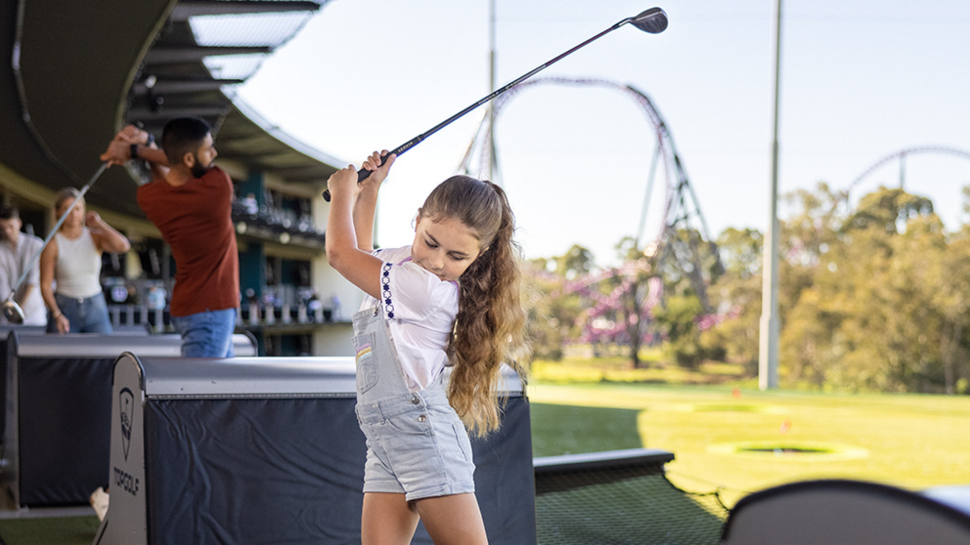 A young girl with long hair swings a golf club at a driving range, while an adult in the background practices their swing. Amusement park roller coasters are visible in the distance.