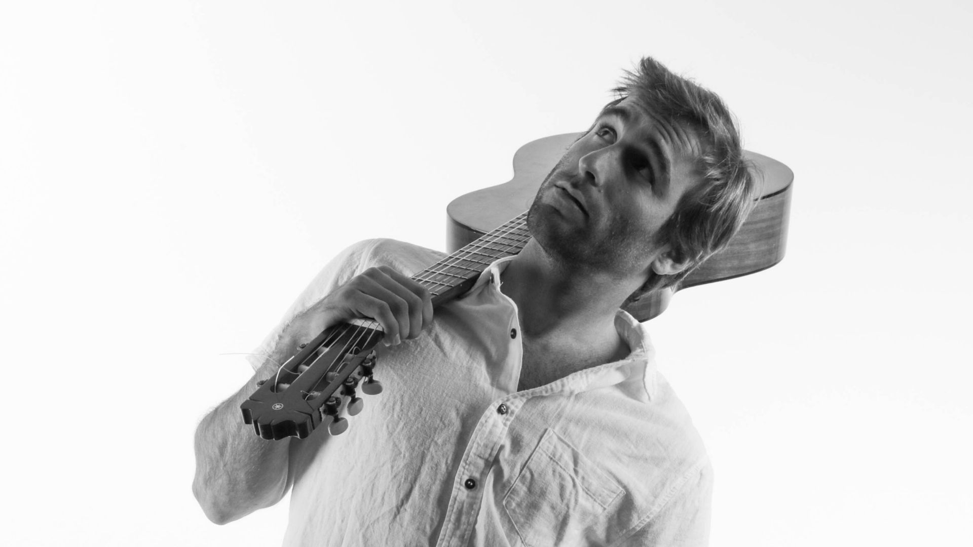 A man wearing a light-colored shirt holds an acoustic guitar over his shoulder and looks upward thoughtfully. The image is in black and white with a plain background.