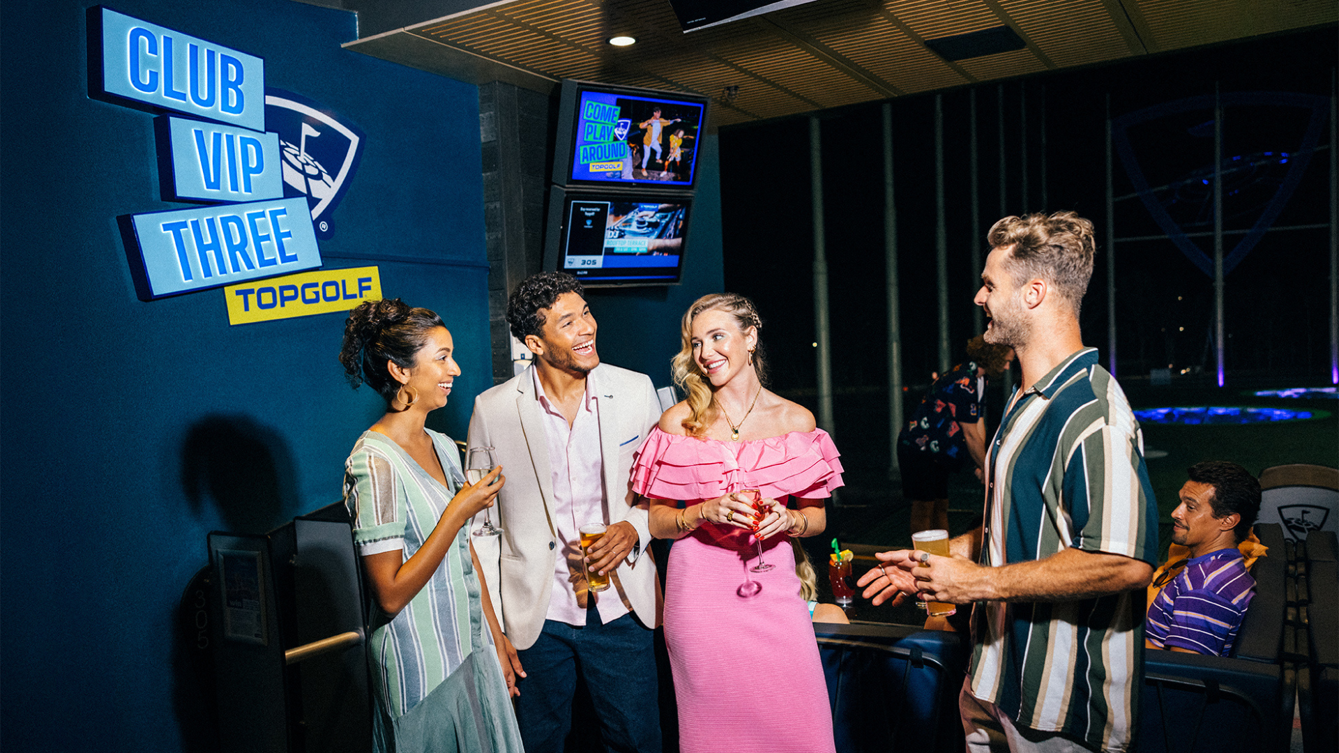 Four people dressed in stylish, semi-formal attire laugh and converse while holding drinks at an indoor golf lounge labeled "Club VIP Three Topgolf." Televisions and a golf range are visible in the background.