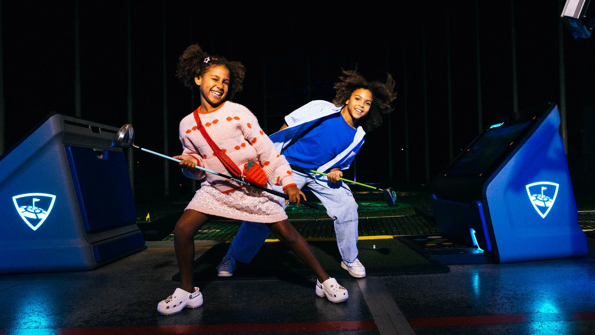 Two kids smiling and posing with golf clubs at an indoor golf entertainment venue, with bright blue-lit golf bays in the background.
