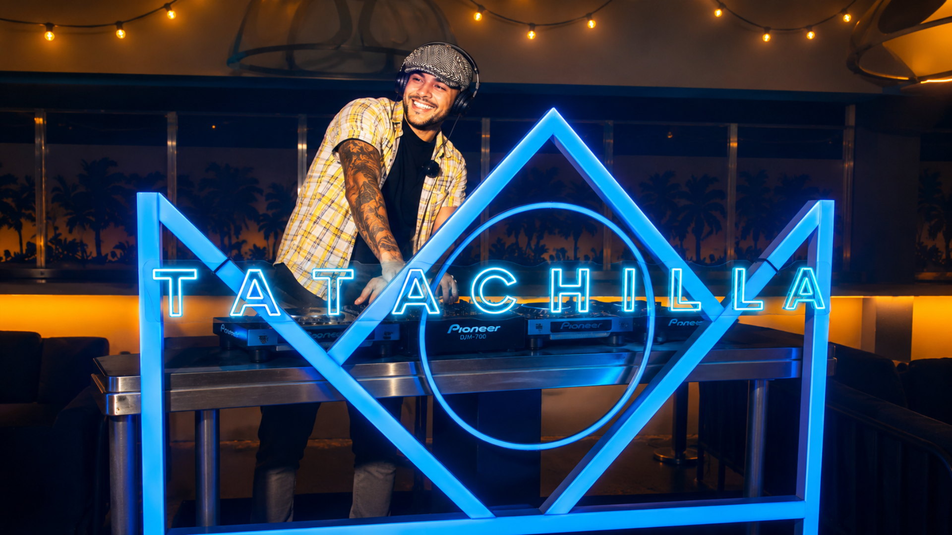 A smiling DJ wearing headphones and a cap stands behind a neon-lit DJ booth that reads "TATACHILLA," with warm glowing lights and palm trees in the background.