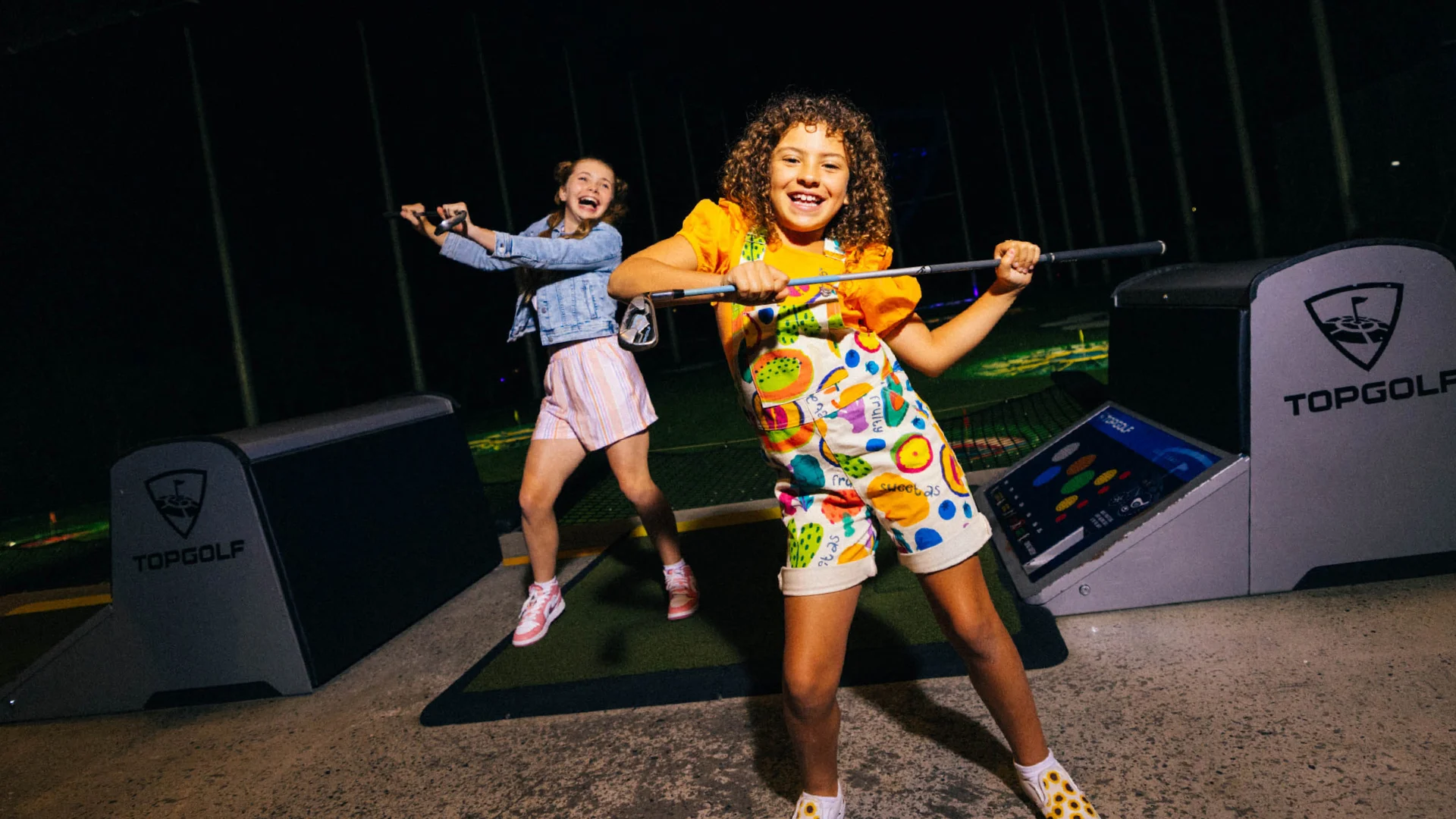 Two kids joyfully swinging golf clubs at an indoor Topgolf venue, with colorful outfits and gaming screens behind them.