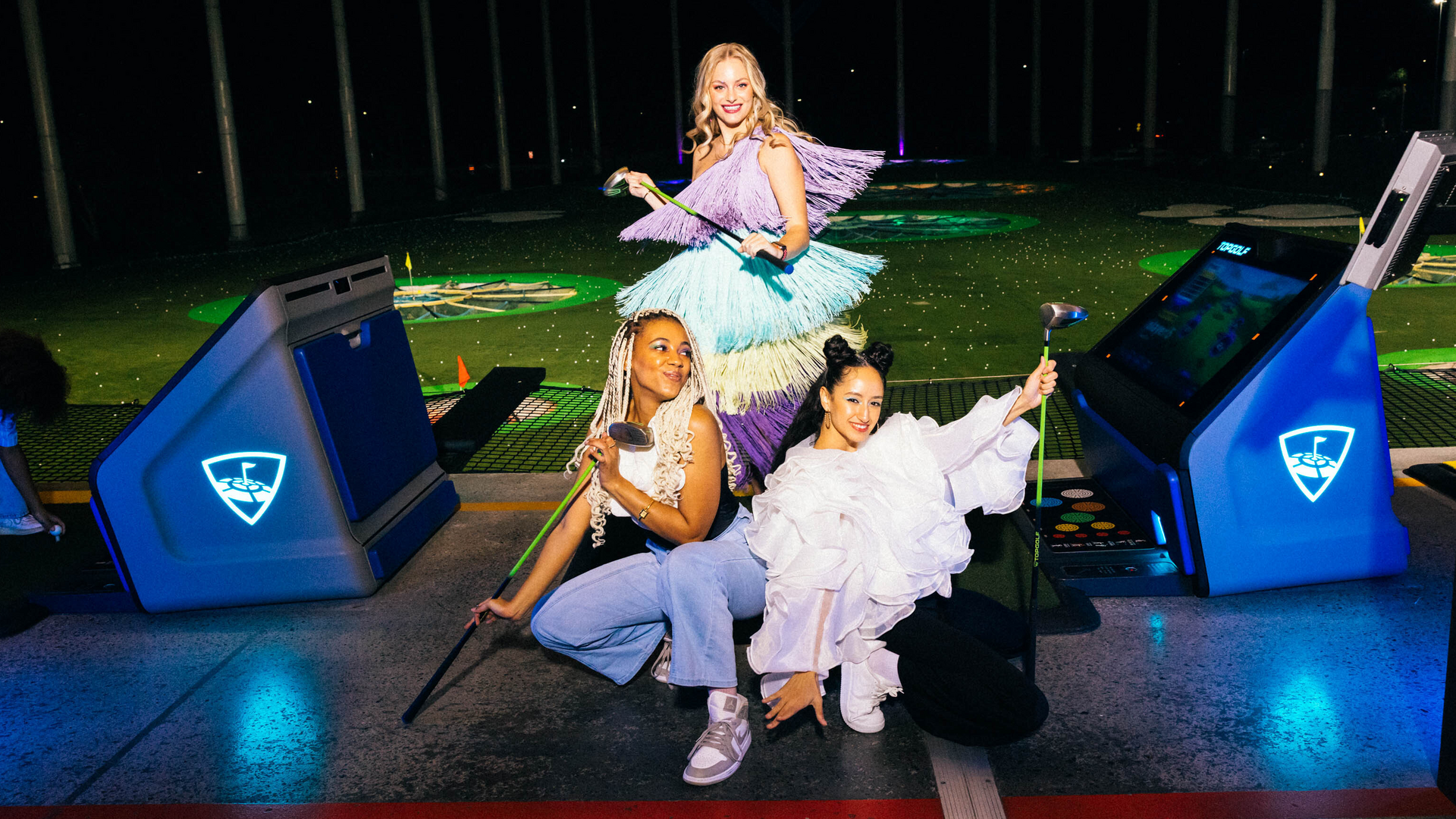 Three women posing with golf clubs at a driving range, wearing colorful and stylish outfits. Two kneel while one stands behind them. The background features an illuminated outdoor golf range.