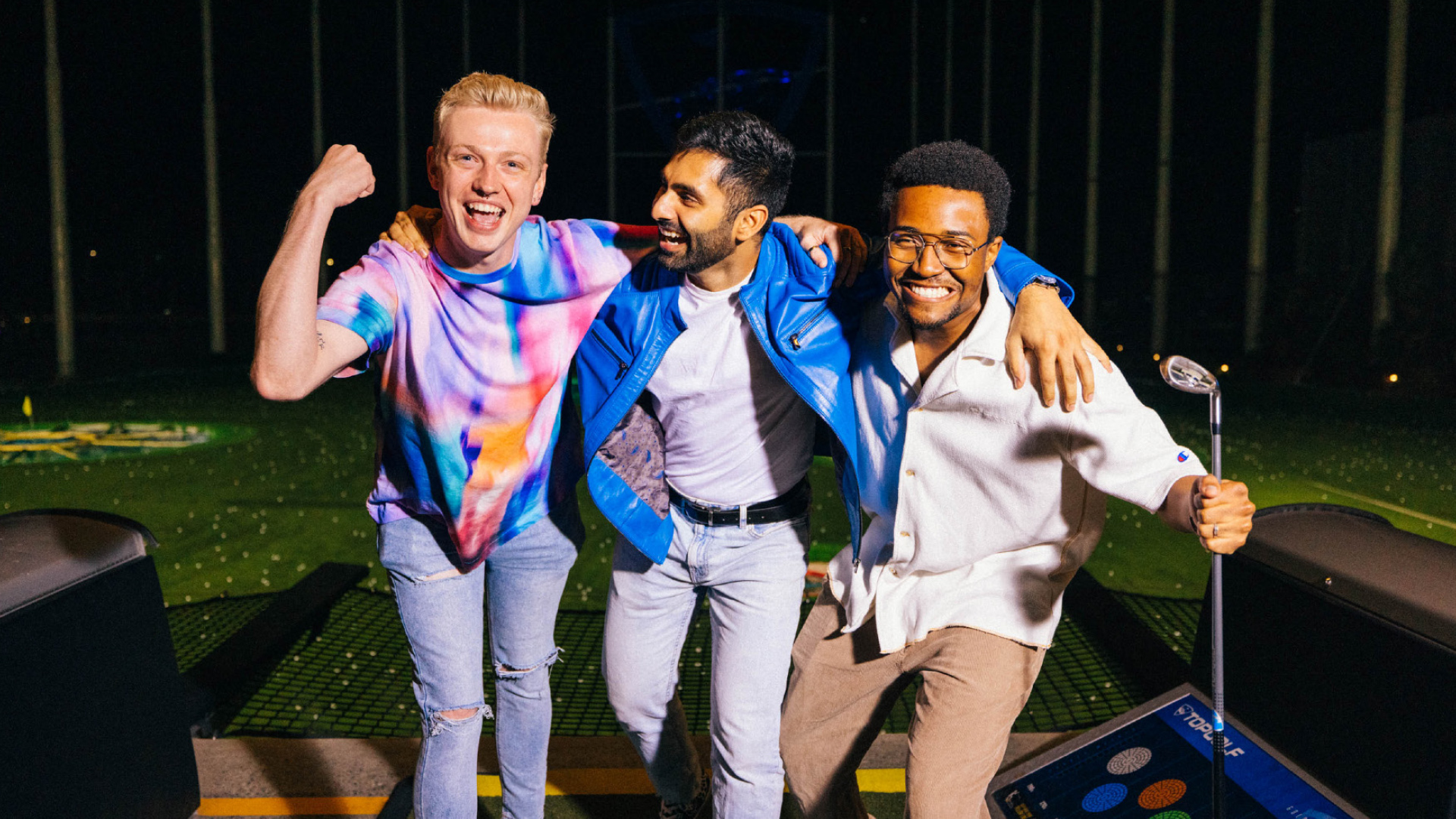 Three friends smile and pose together at a driving range at night; one flexes his arm, another stands with arms around friends, and the third holds a golf club. They look happy and excited.