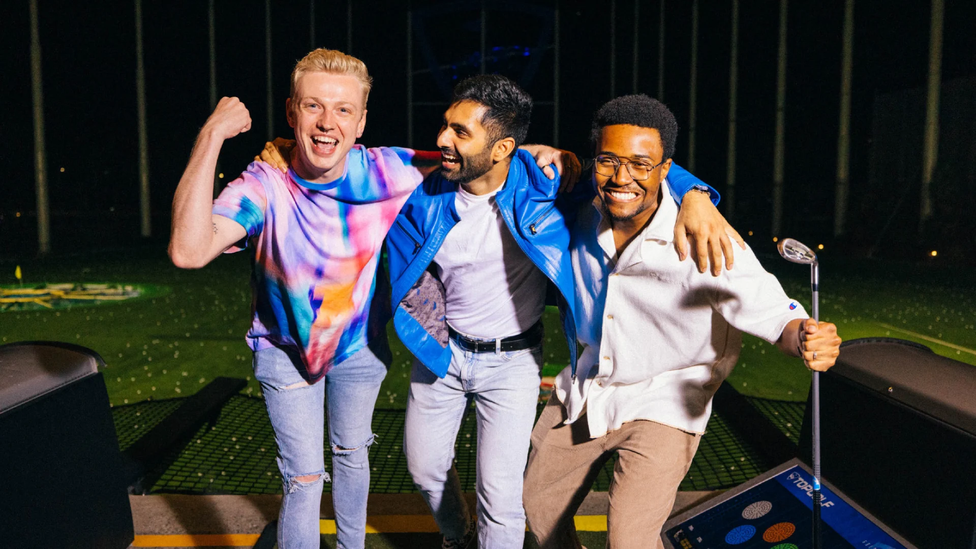 Three friends smile and pose together at a driving range at night; one flexes his arm, another stands with arms around friends, and the third holds a golf club. They look happy and excited.