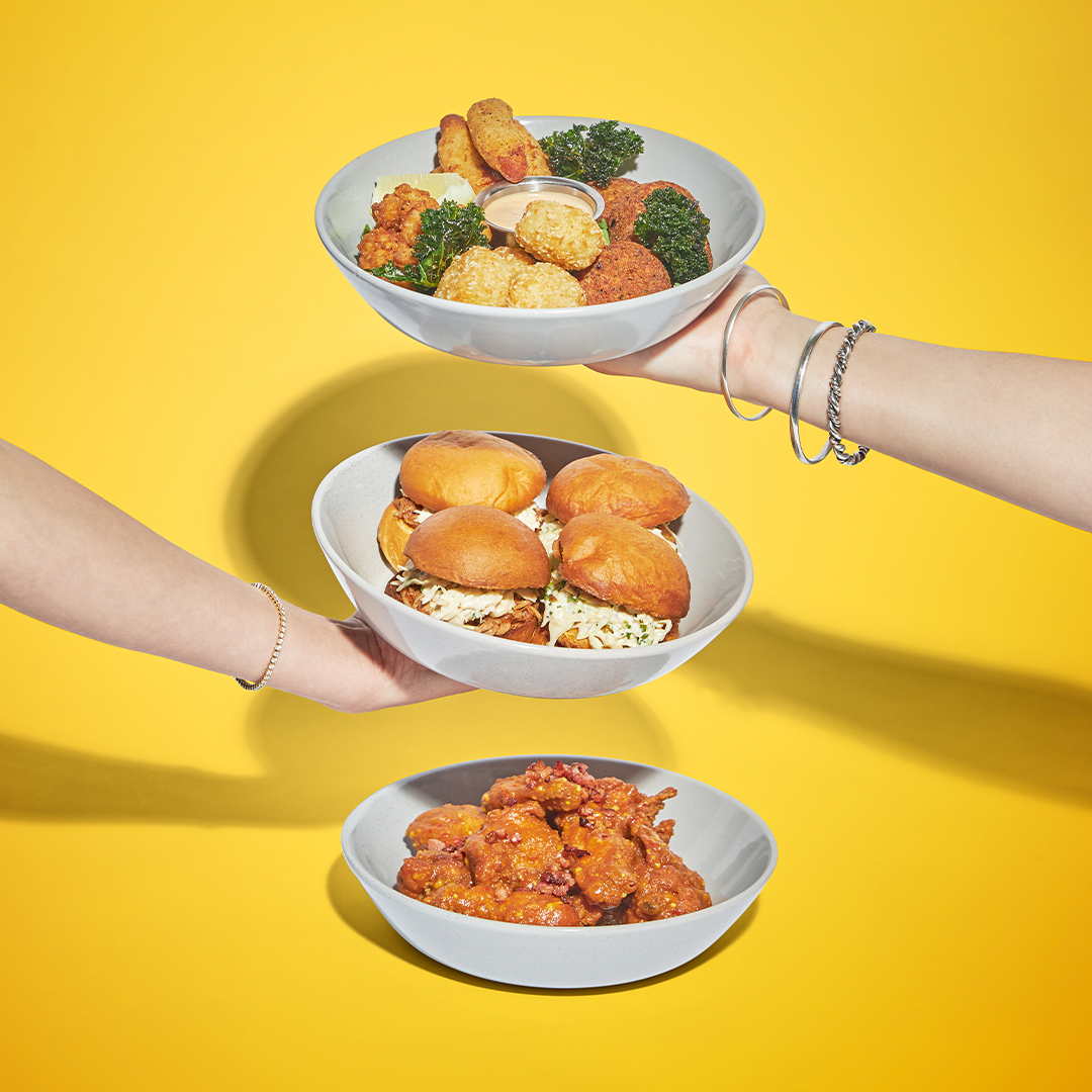 Three hands hold bowls stacked vertically against a yellow background. The top bowl has fried snacks, the middle has sliders, and the bottom bowl contains saucy chicken wings.