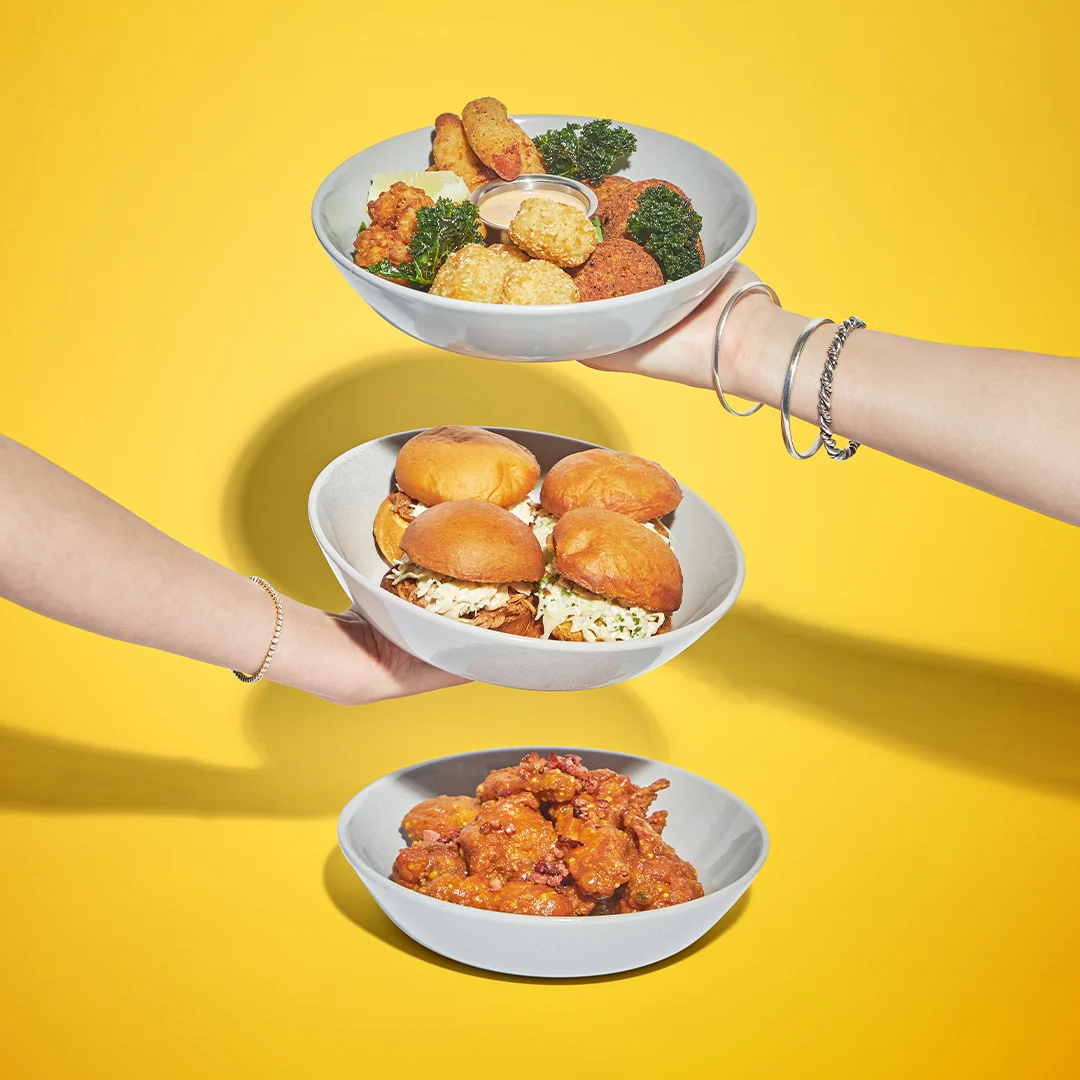 Three hands hold bowls stacked vertically against a yellow background. The top bowl has fried snacks, the middle has sliders, and the bottom bowl contains saucy chicken wings.