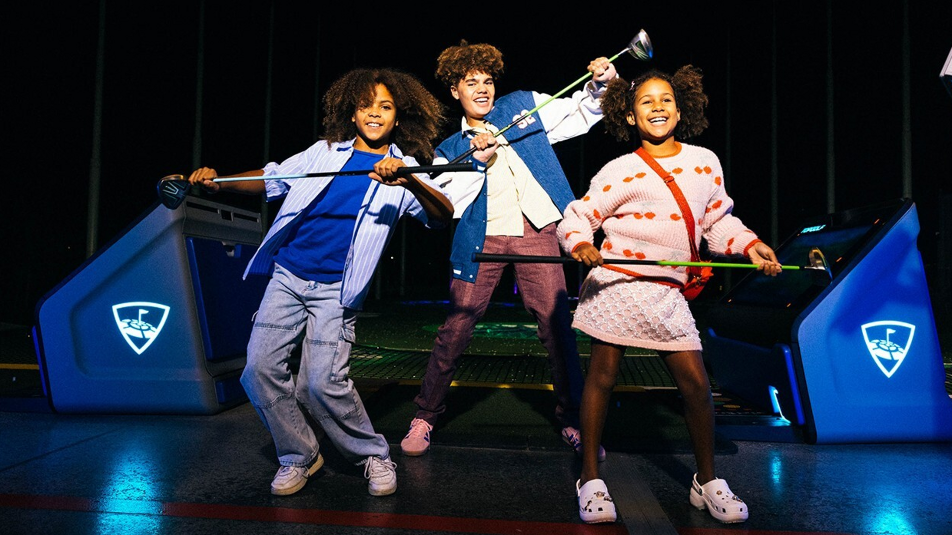 Three smiling kids stand indoors, each holding a golf club and posing playfully at a golf entertainment venue, surrounded by blue-lit golf bays.
