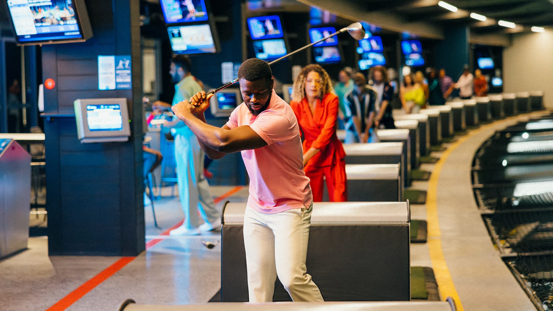 A man in a pink shirt swings a golf club at an indoor driving range, with several people watching and waiting their turn in the background. Multiple TV screens and golf bays are visible.