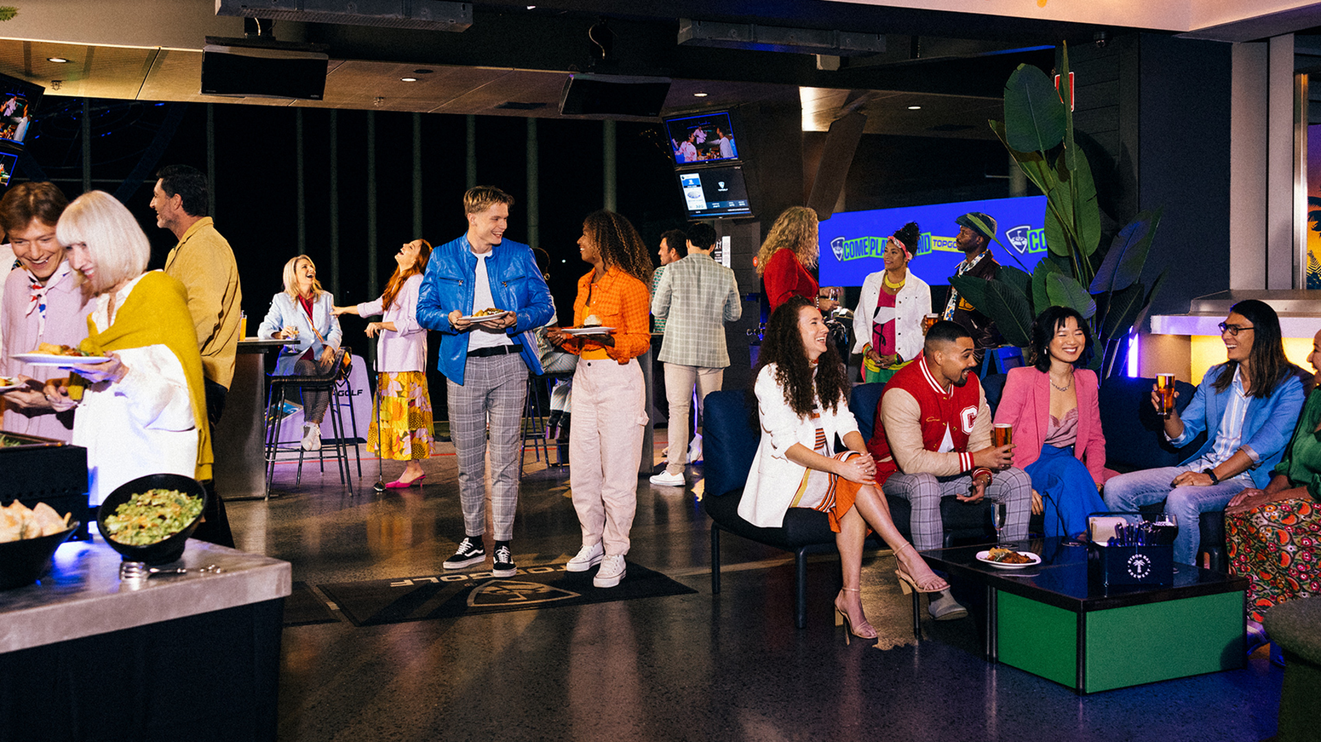 A group of people socialize, eat, and drink at a lively indoor event space with colorful decor, food trays, and lounge seating. Some people are standing and chatting, while others are seated and raising glasses.
