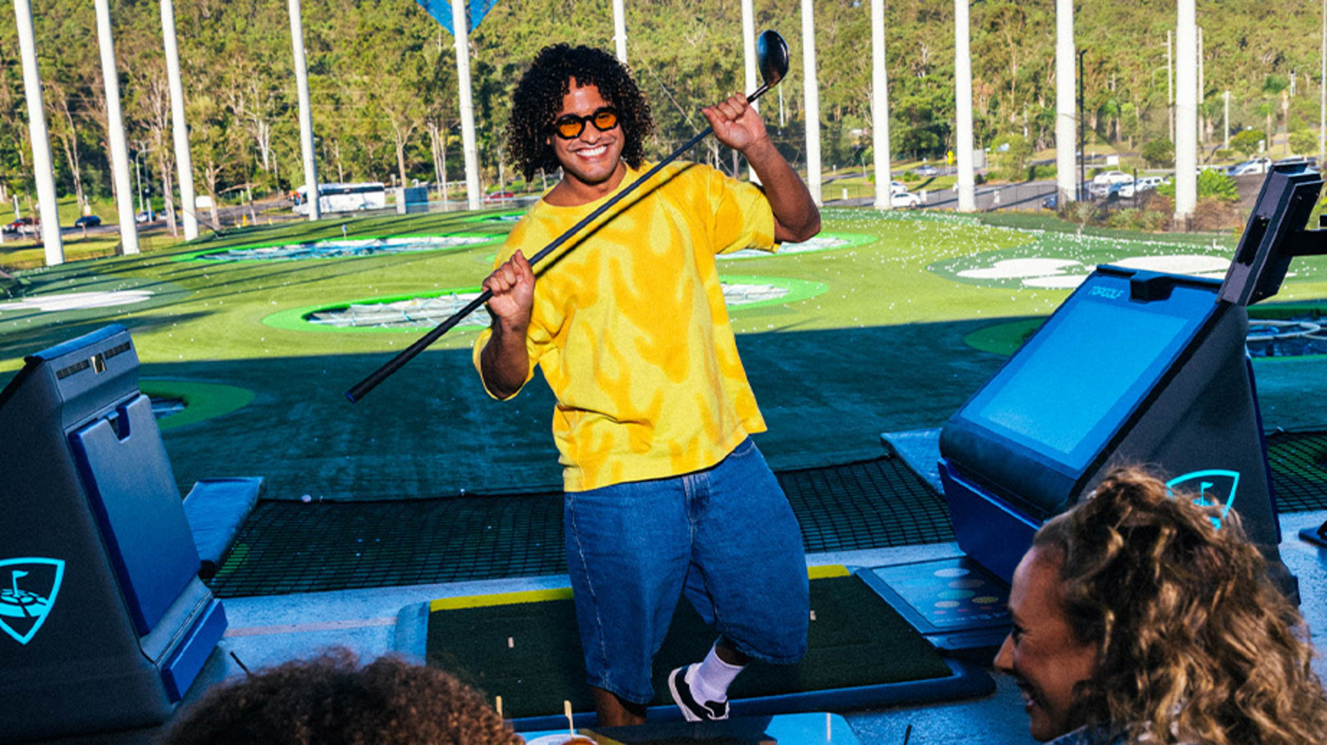 A person wearing a yellow shirt, sunglasses, and shorts smiles and poses with a golf club at an outdoor driving range, while others watch and laugh in the foreground.