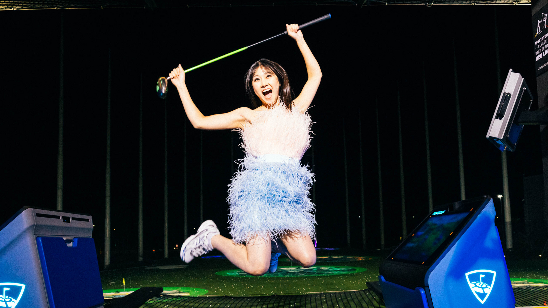 A woman in a feathered dress jumps joyfully while holding a golf club at an outdoor driving range at night.