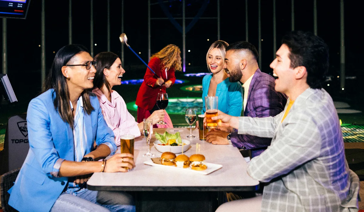 A group of young adults laughs and enjoys drinks and sliders at an outdoor golf entertainment venue at night, with one person swinging a golf club in the background.