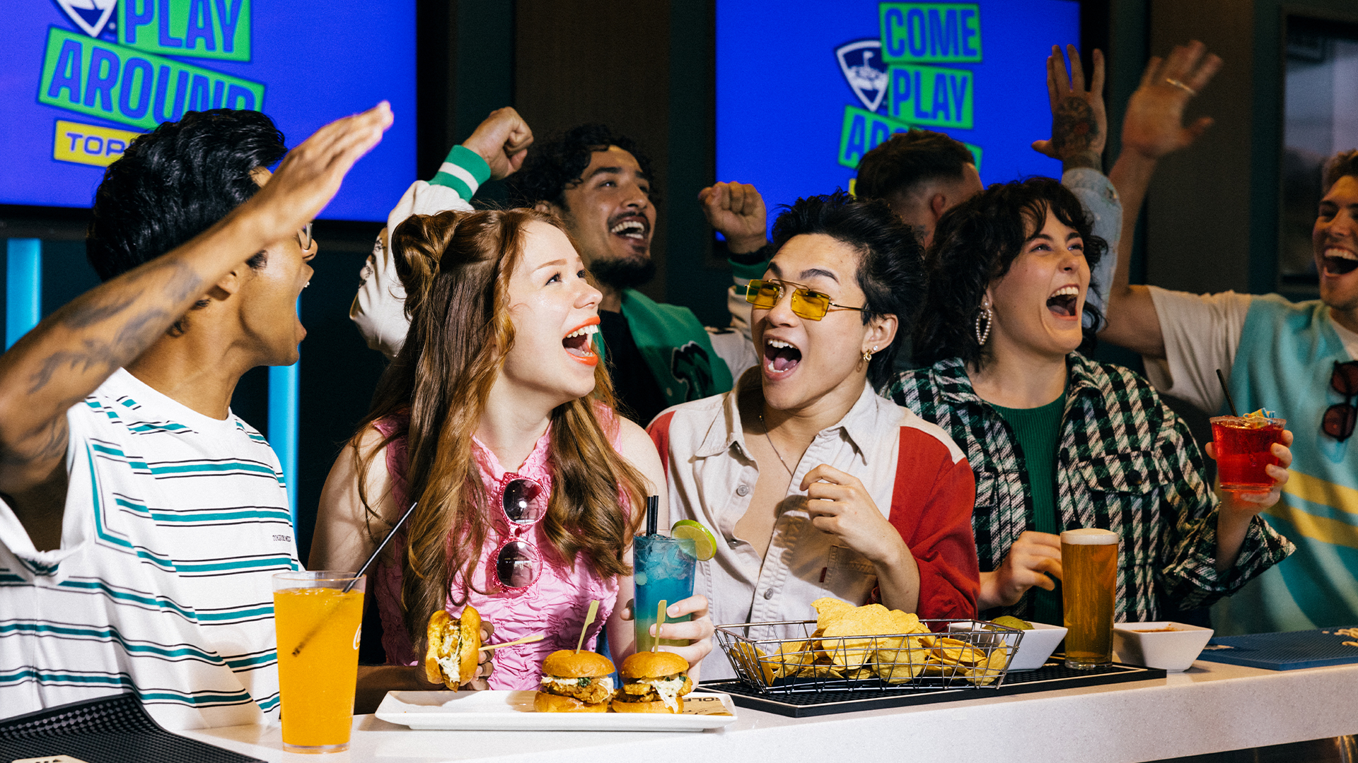 A group of young adults sit at a bar, laughing and cheering with drinks and food in front of them. In the background, screens display the words “Come Play Around.” The atmosphere is lively and festive.