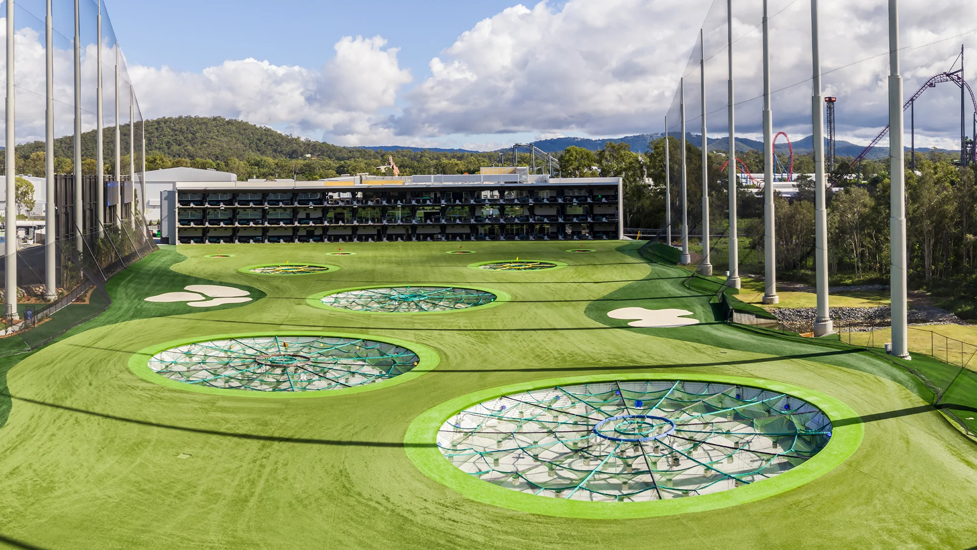 A modern outdoor golf driving range with multiple levels of hitting bays overlooks a bright green field featuring large, circular targets and surrounding netting under a partly cloudy sky.