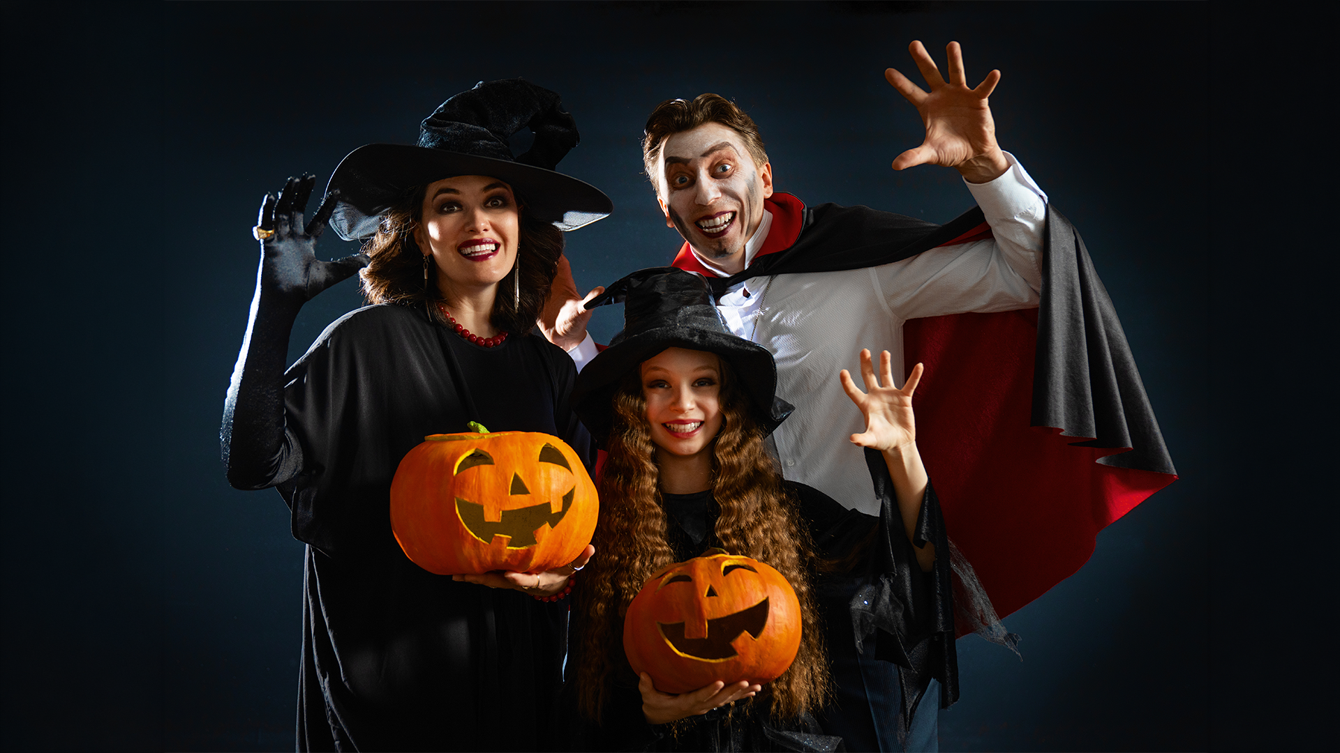 A woman dressed as a witch, a man dressed as a vampire, and a girl dressed as a witch pose for a Halloween photo while holding jack-o’-lanterns.