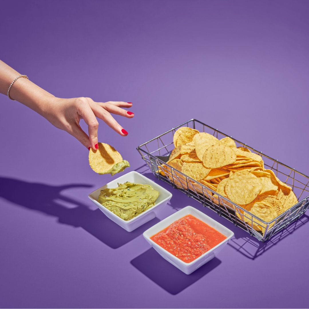 A hand with red nails dips a tortilla chip into guacamole beside a basket of chips and a bowl of salsa, all on a purple background.