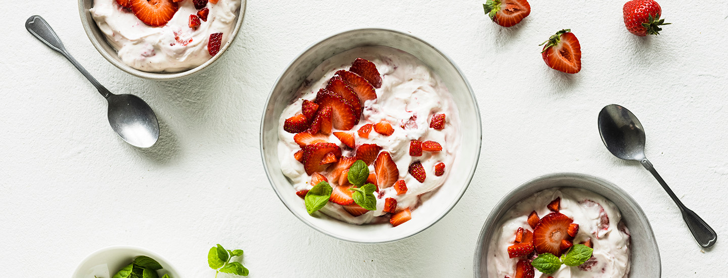 Mansikkarahkaa valkoisessa kulhossa. Rahkan päälle on aseteltu mansikkaviipaleita ja tuoreita mintunlehtiä. Kulhon ylä- ja alapuolella on kaksi pienempää kulhoa. K-Ruoka välipalaresepti.