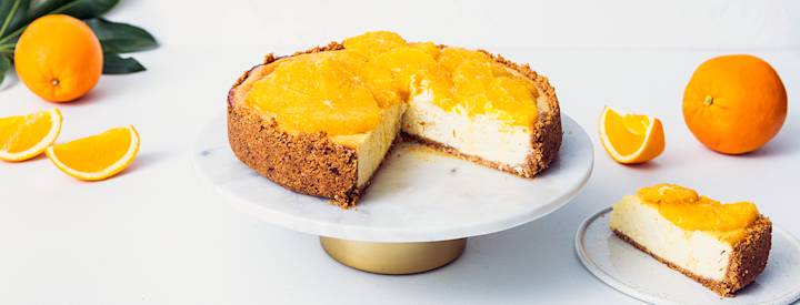 Paistettu appelsiinijuustokakku vaalealla kakkulautasella. Kakkulautasen ympärillä pöydällä on kokonaisia appelsiineja ja leikattuja appelsiinilohkoja. Kakkulautasen vieressä oikealla puolella on pieni lautanen, jolla on siivu appelsiinijuustokakkua. K-Ruoka juustokakkuresepti.