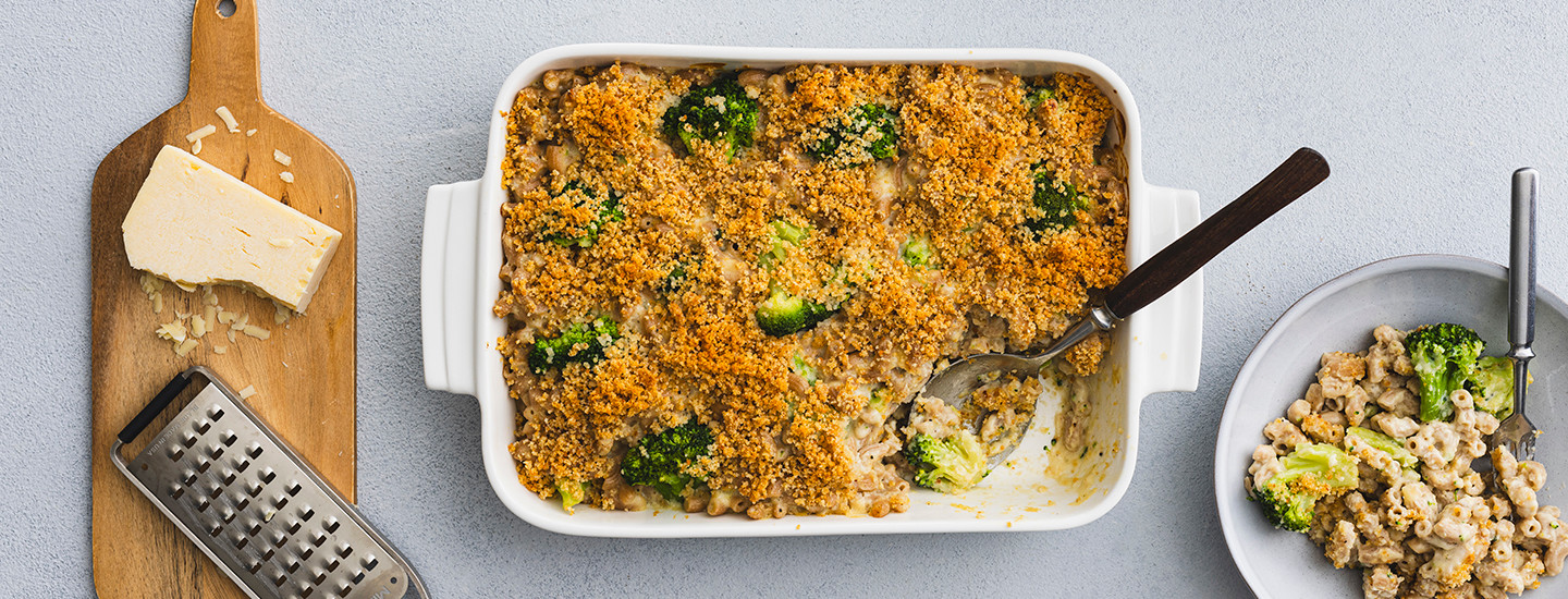 Makaronilaatikkotyylinen parsakaali mac 'n' cheese valkoisessa vuoassa, vieressä lautanen, jossa parsakaali mac 'n' cheeseä sekä leikkuulauta, jonka päällä pala cheddarjuustoa.